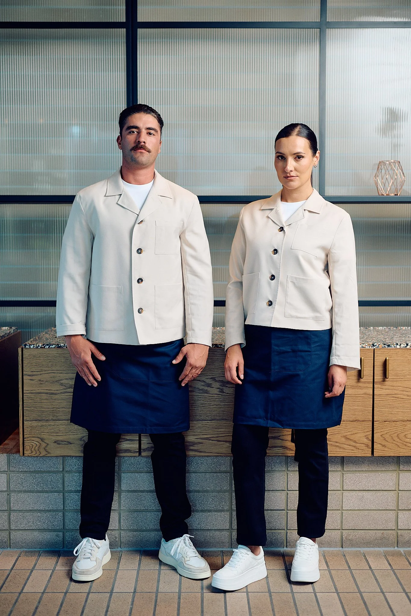 Two people standing indoors in front of a glass wall, wearing white chef jackets and navy aprons, looking at the camera.