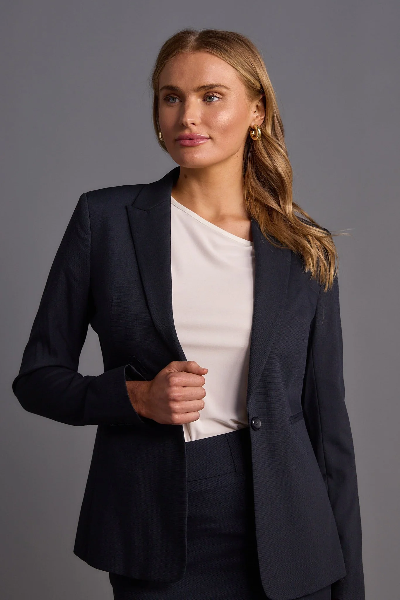 A woman with blonde hair in a deep side part, wearing a black blazer over a white top, and gold hoop earrings, standing against a plain gray background.