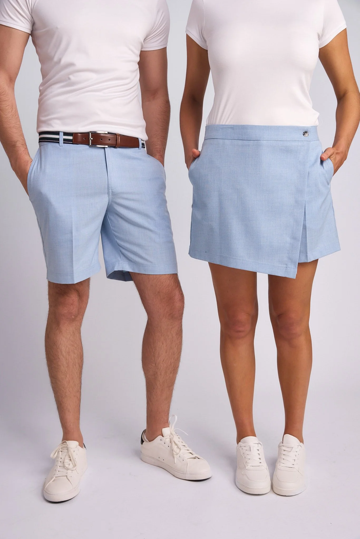 A man and woman wearing white t-shirts, light blue shorts and skirt, and white sneakers posing against a plain background.