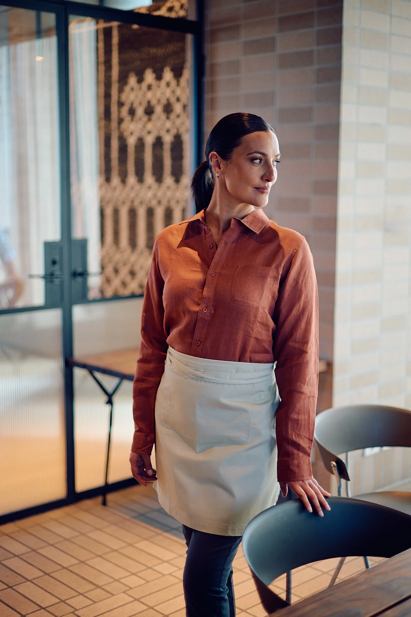 A woman with dark hair tied back, wearing a burnt orange long-sleeve button-up shirt and a white apron, standing in a modern cafe or restaurant with a brick wall and a glass partition in the background.