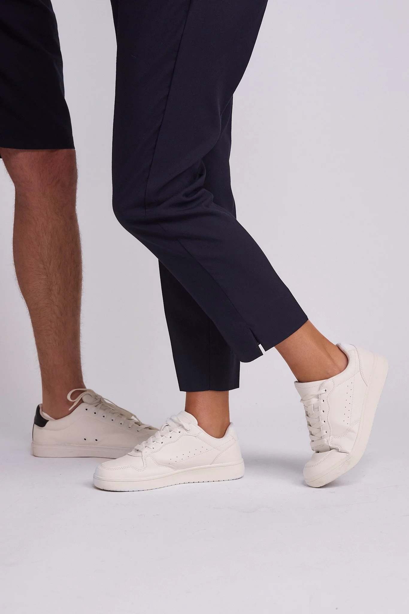 Close-up of two people standing on a white background. One person is wearing dark pants and white sneakers, with one foot slightly raised. The other person is wearing shorts and white sneakers, with one foot flat on the ground.