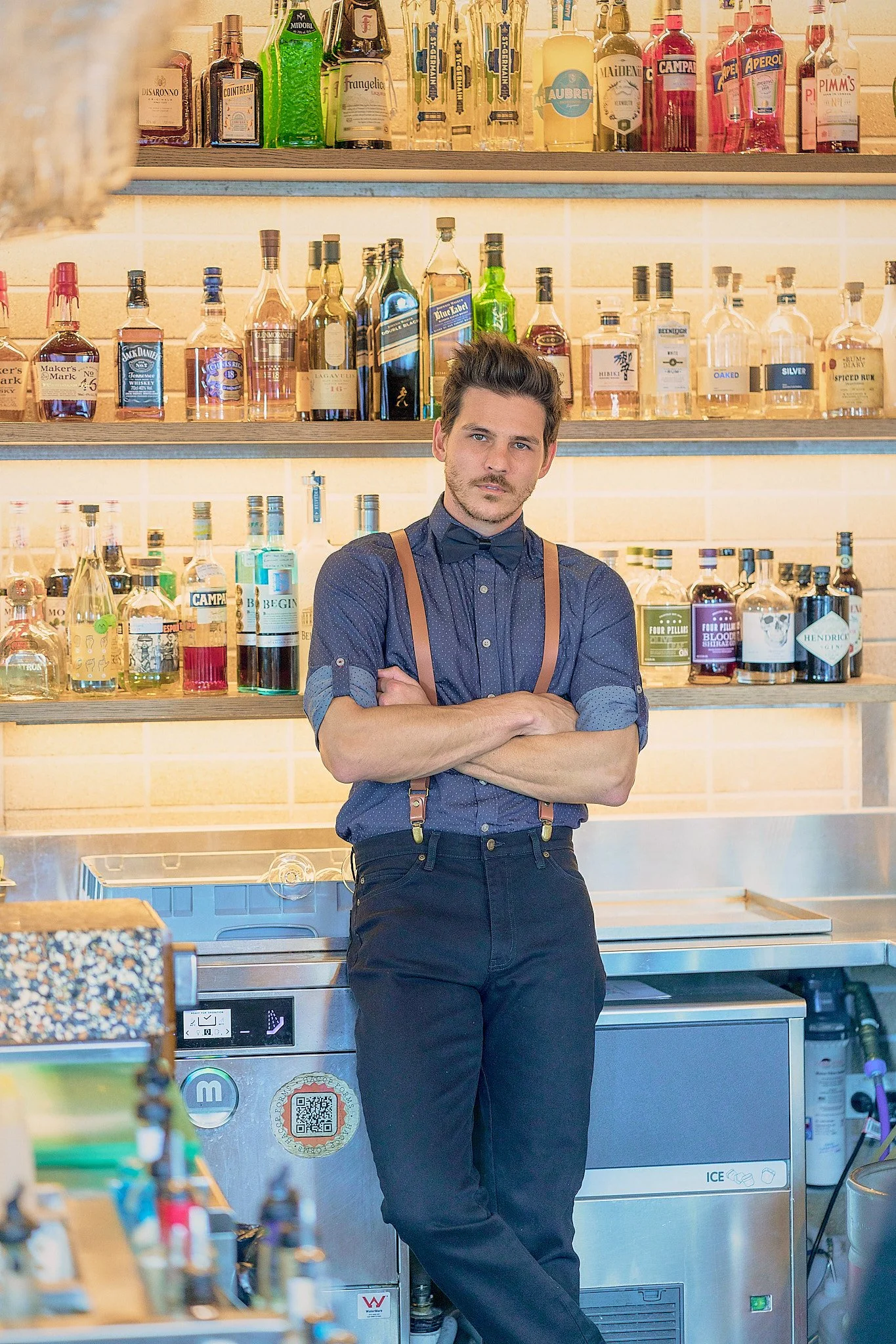 A man with dark hair and a mustache in a bartender's uniform with suspenders, standing with crossed arms behind a bar with many bottles of alcohol on shelves behind him.