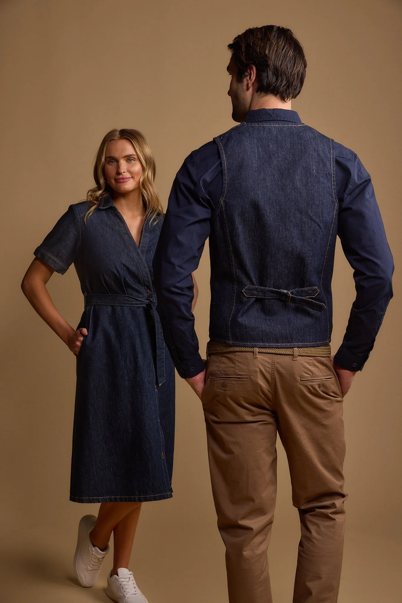 A woman and a man wearing denim outfits, standing against a beige background. The woman has blonde hair and is smiling, dressed in a denim dress with short sleeves and white sneakers. The man has brown hair and is facing away from the camera, wearing