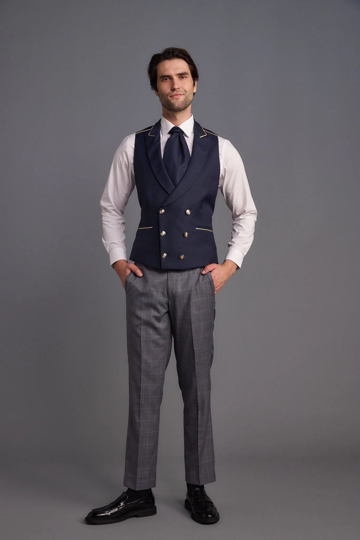 A man in a double-breasted navy vest, white shirt, gray checkered pants, and black shoes standing against a plain gray background.