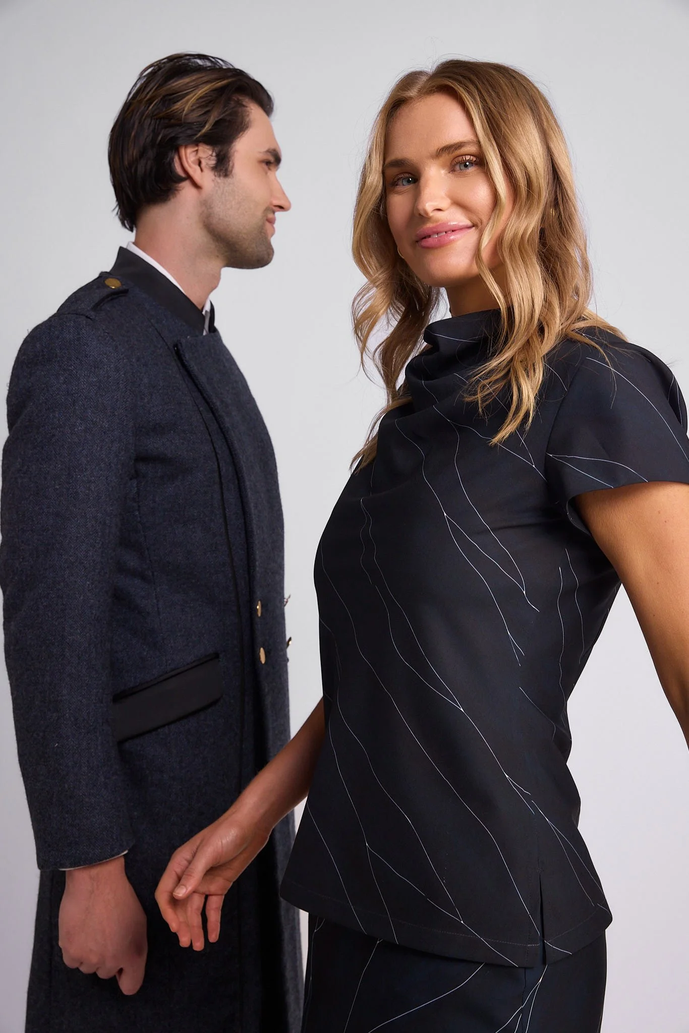 A man and woman standing close together, the woman smiling at the camera, both dressed in dark formal clothing against a plain background.