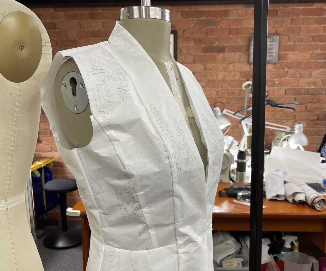 A dress form covered with a pattern paper in a sewing or fashion design workspace, with sewing supplies and lamps on a desk in the background.