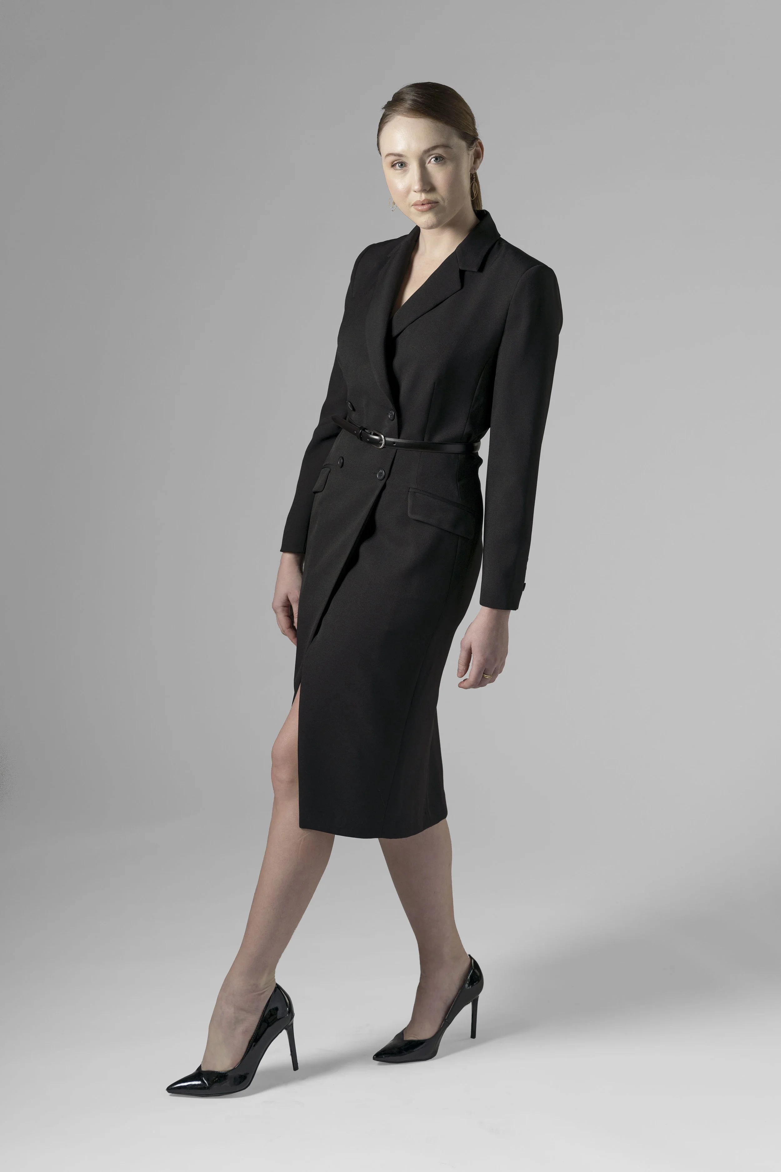 A woman in a black blazer dress with high heels standing against a gray background.