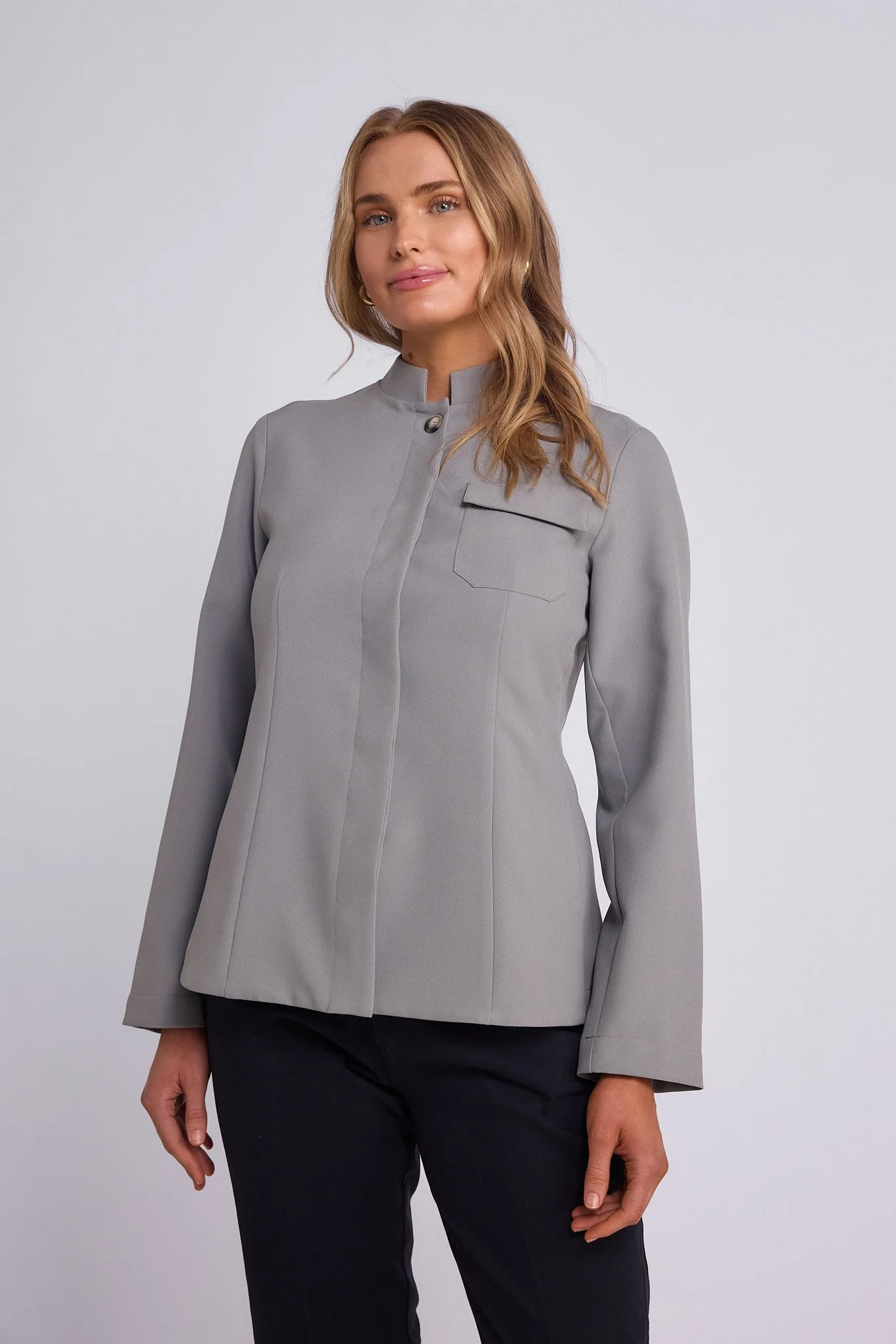 A woman with long wavy blonde hair wearing a light grey tailored blazer with a front pocket, standing against a plain light grey background.