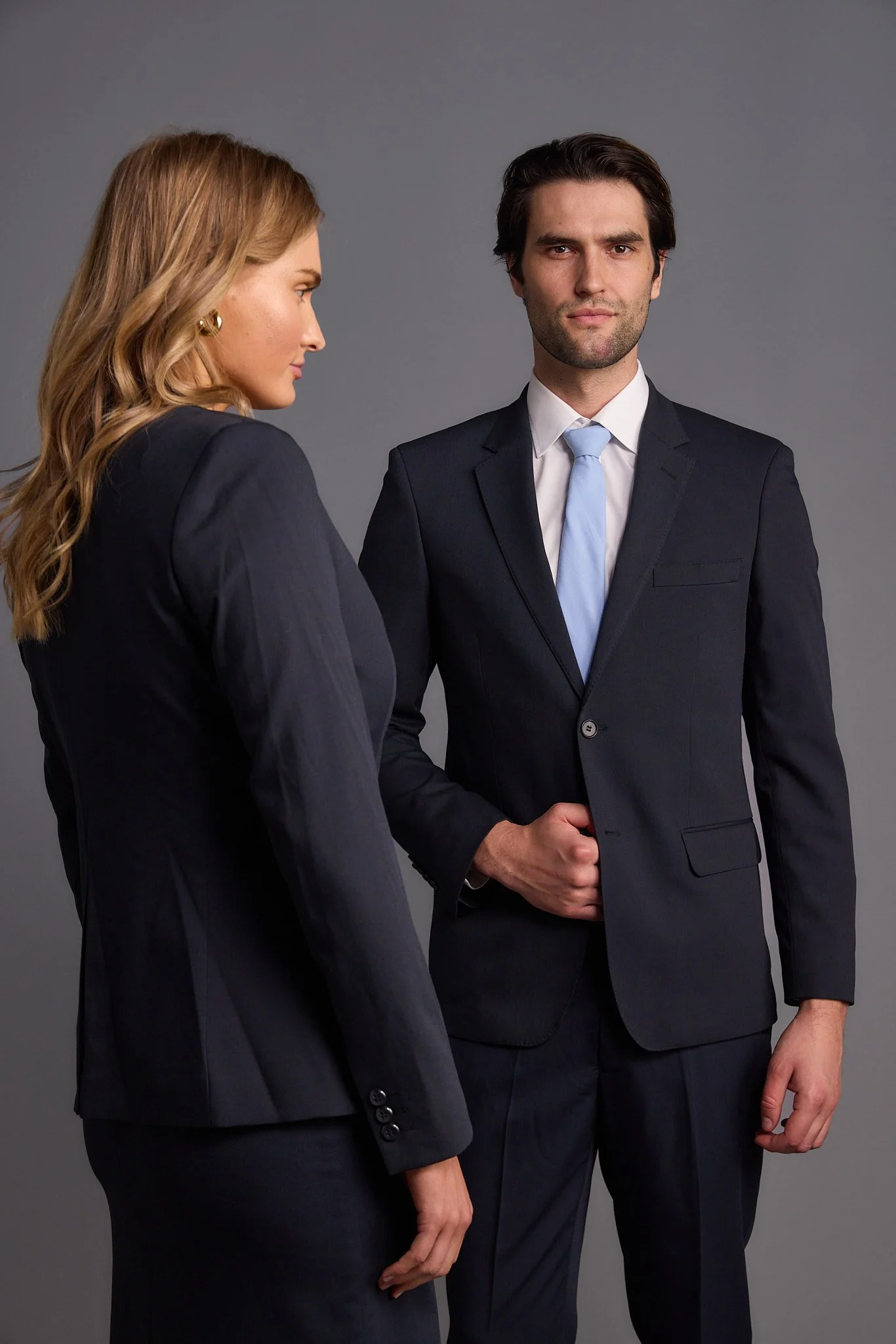 A woman with wavy blonde hair and earrings looking at a man with dark hair, both dressed in black business suits, against a plain gray background.