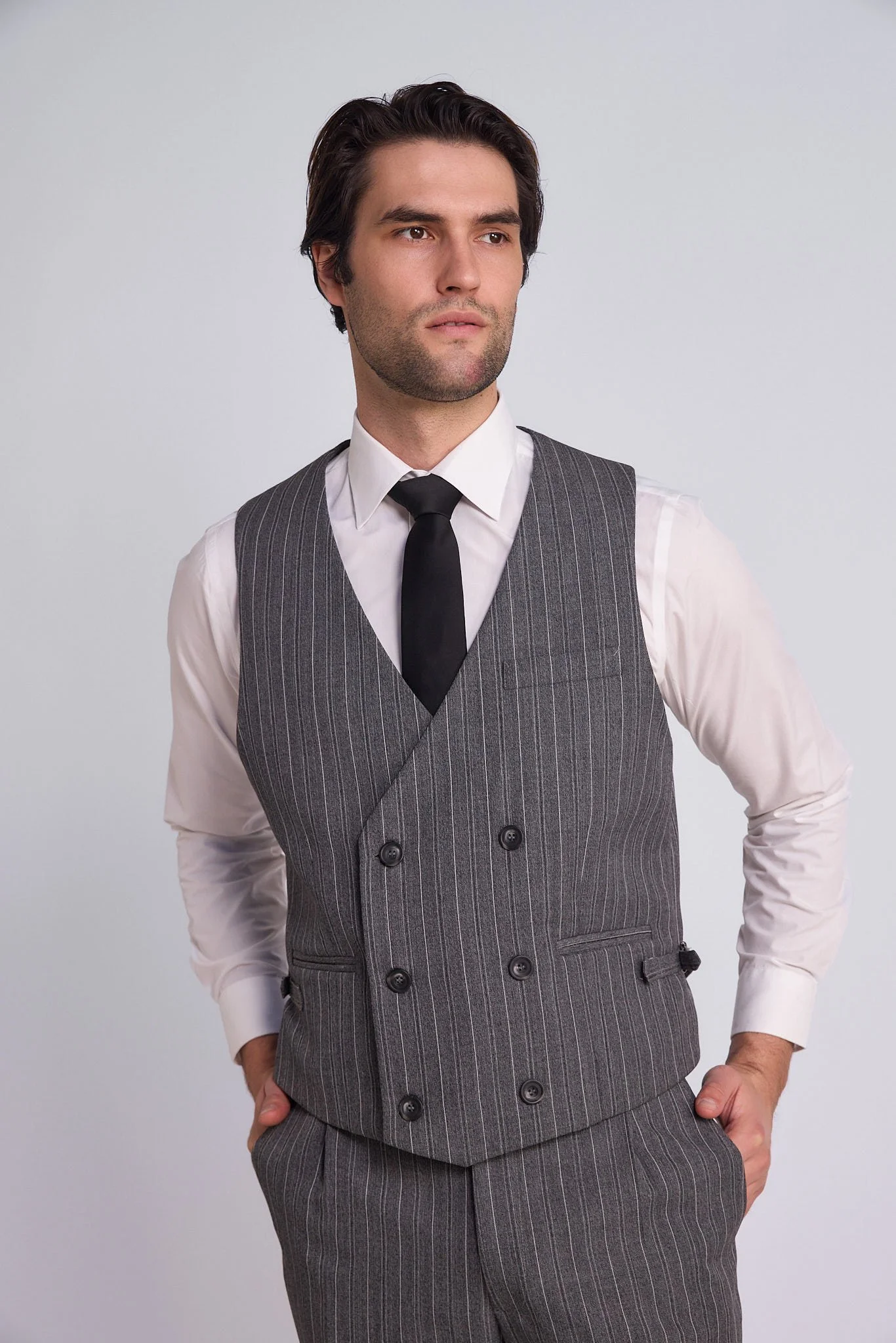 A man wearing a white shirt, black tie, and gray striped vest with a matching pair of pants, standing against a plain white background.