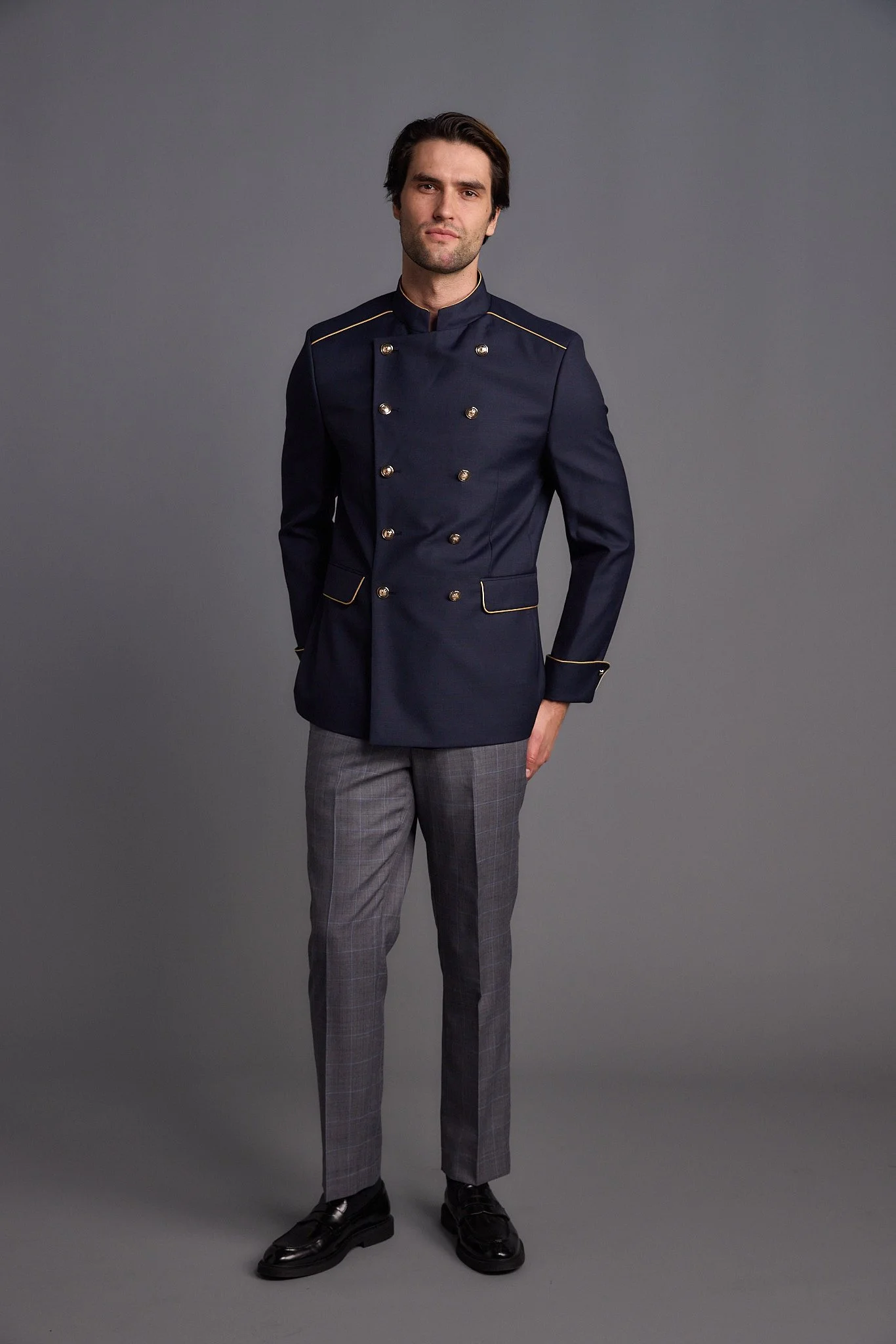 Man wearing a dark blue military-style jacket with gold buttons and piping, gray checkered pants, and black shoes, standing against a gray background.