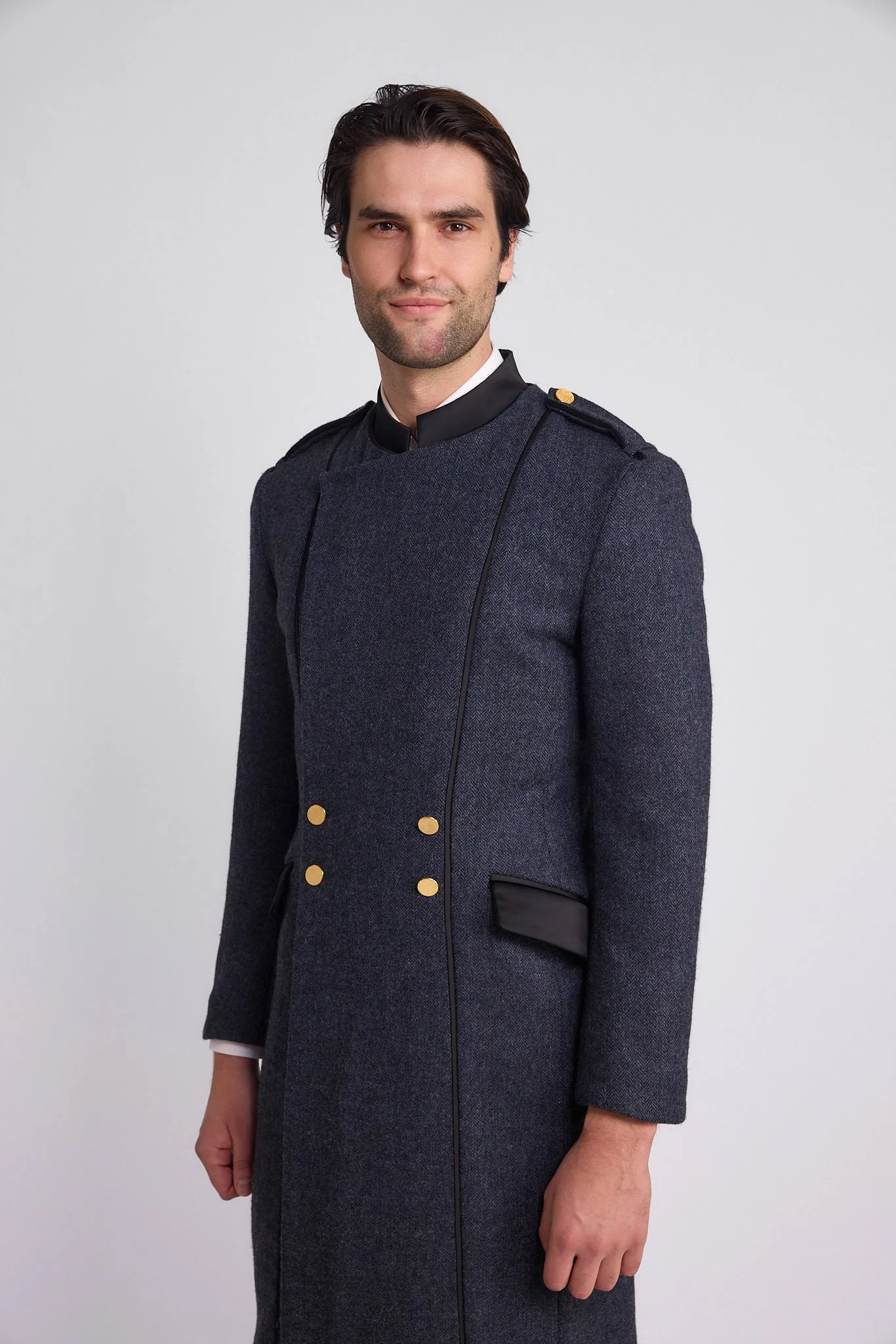 A man in a dark gray military-style coat with gold buttons and shoulder epaulets, standing against a plain light gray background.