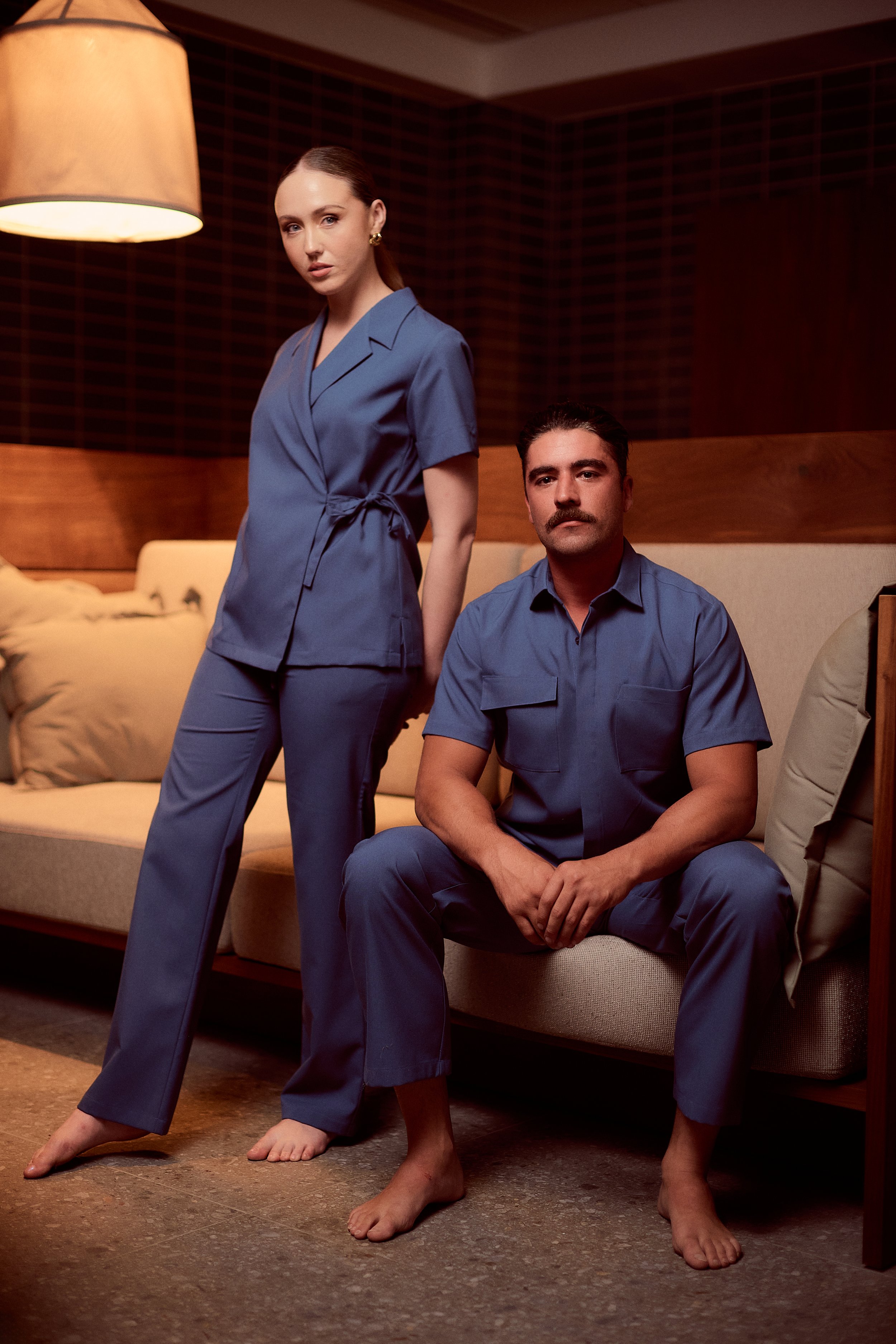 A woman and a man in blue pajamas indoors, with the woman standing and the man sitting on a beige couch.