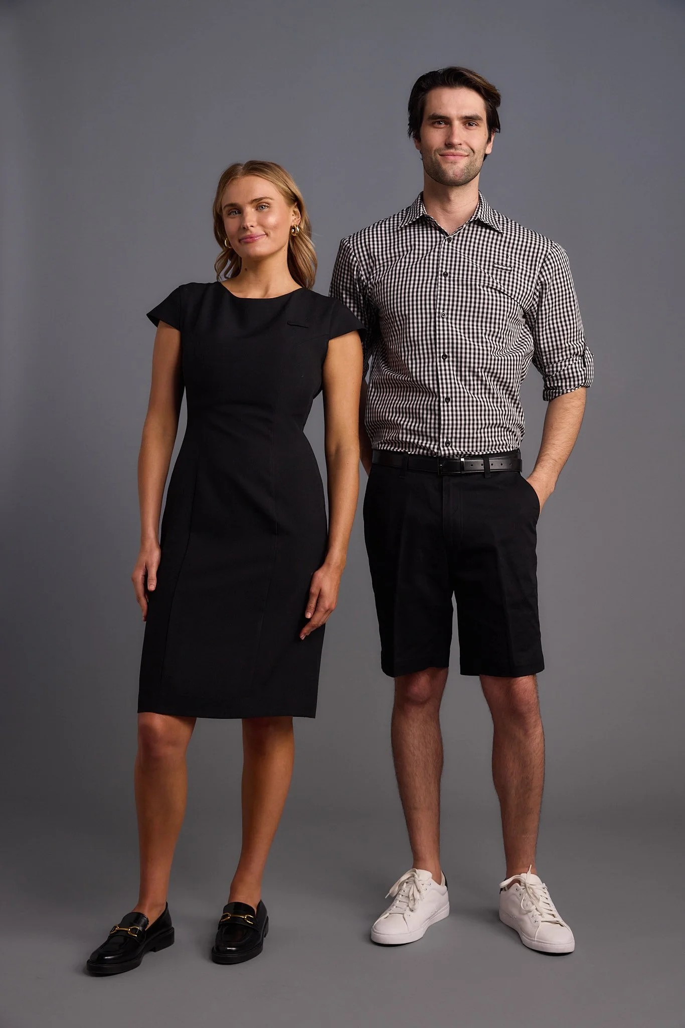 A woman in a black dress and a man in a checkered shirt and shorts standing against a plain gray background.