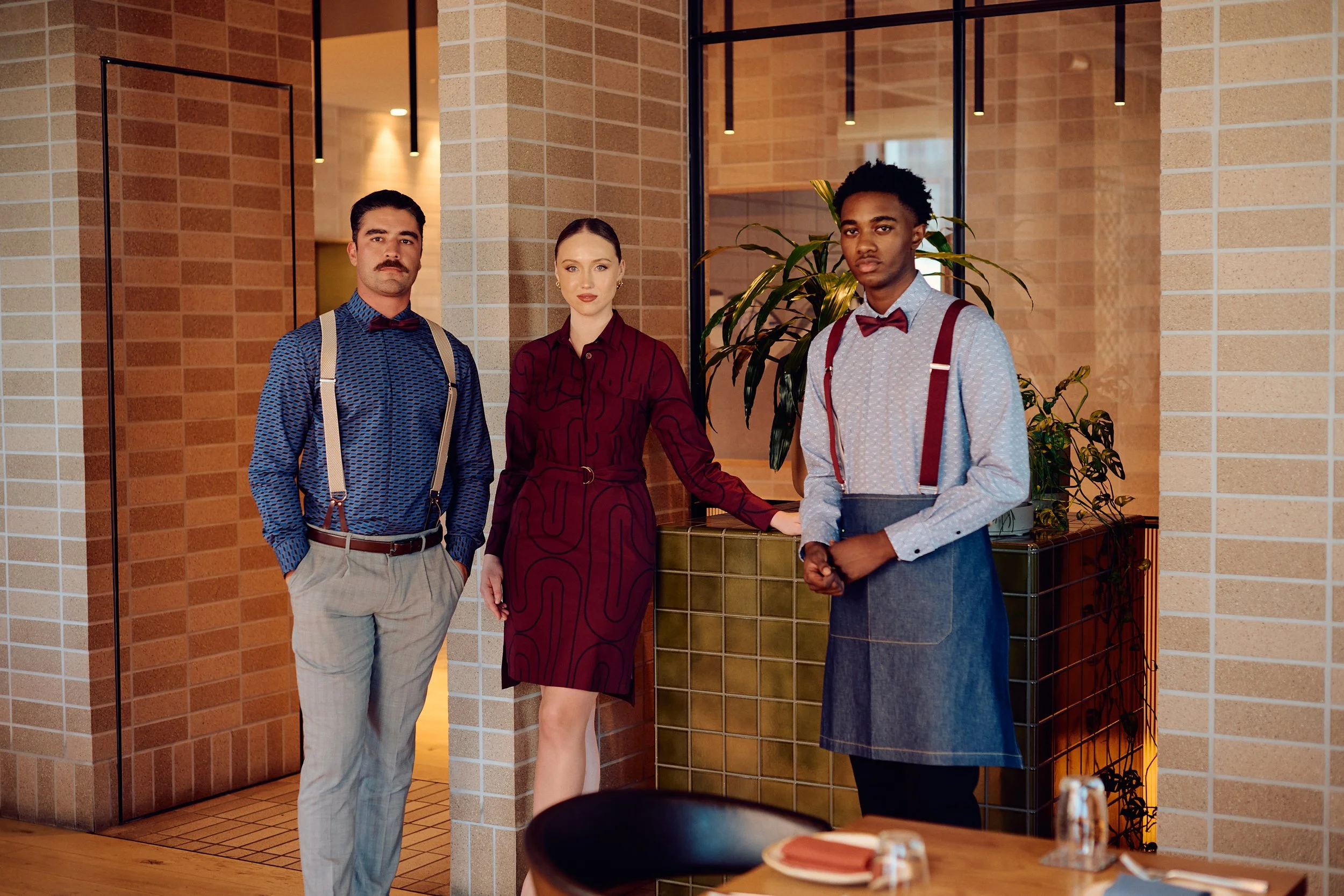 Three waitstaff members stand inside a restaurant near a wall with brick and glass elements. The man on the left wears a blue shirt with suspenders, a red bow tie, and light-colored trousers. The woman in the middle wears a burgundy dress with a belt. The man on the right wears a light-colored shirt, red suspenders, a bow tie, and an apron. There is a table with silverware and place settings in the foreground and a large plant behind the staff.