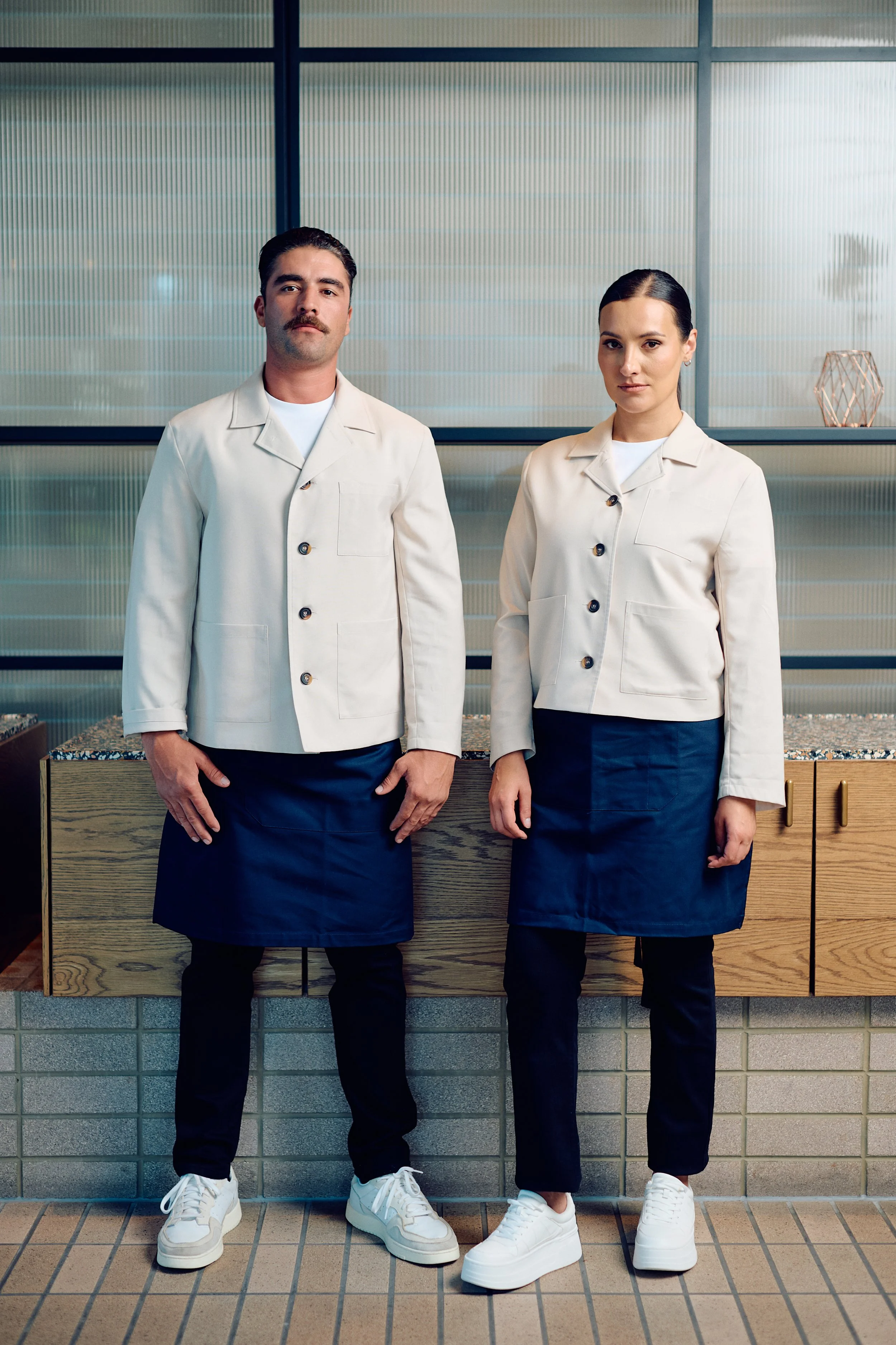 Two menu staff members, a man and a woman, standing side by side indoors, wearing white jackets, navy aprons, black pants, and white shoes, in front of a textured wall and wooden counter.