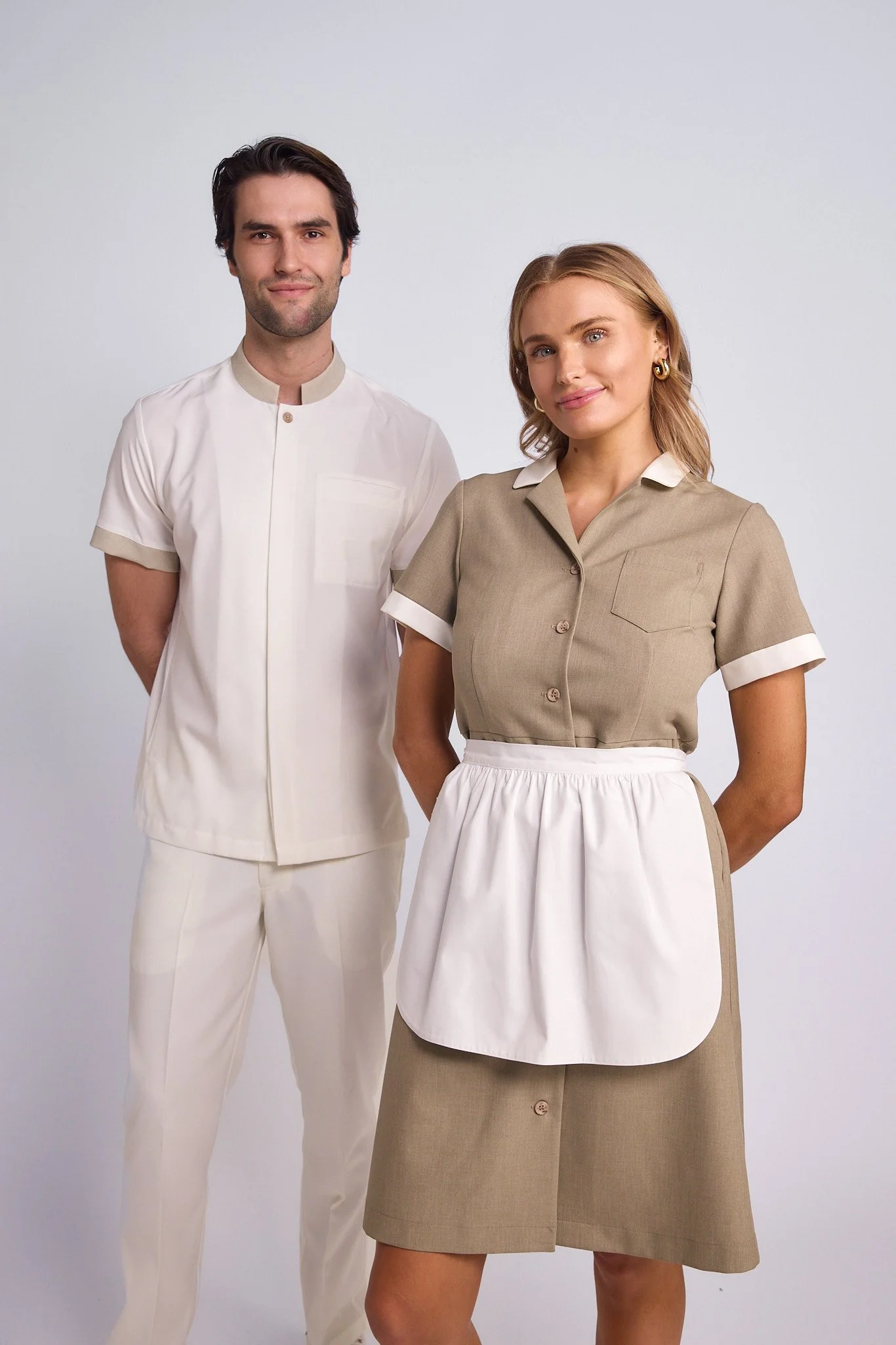 A man and woman dressed in vintage-style uniforms, standing against a plain background. The man wears a white short-sleeved shirt and matching pants. The woman wears a beige dress with white accents and an apron. Both are smiling slightly.
