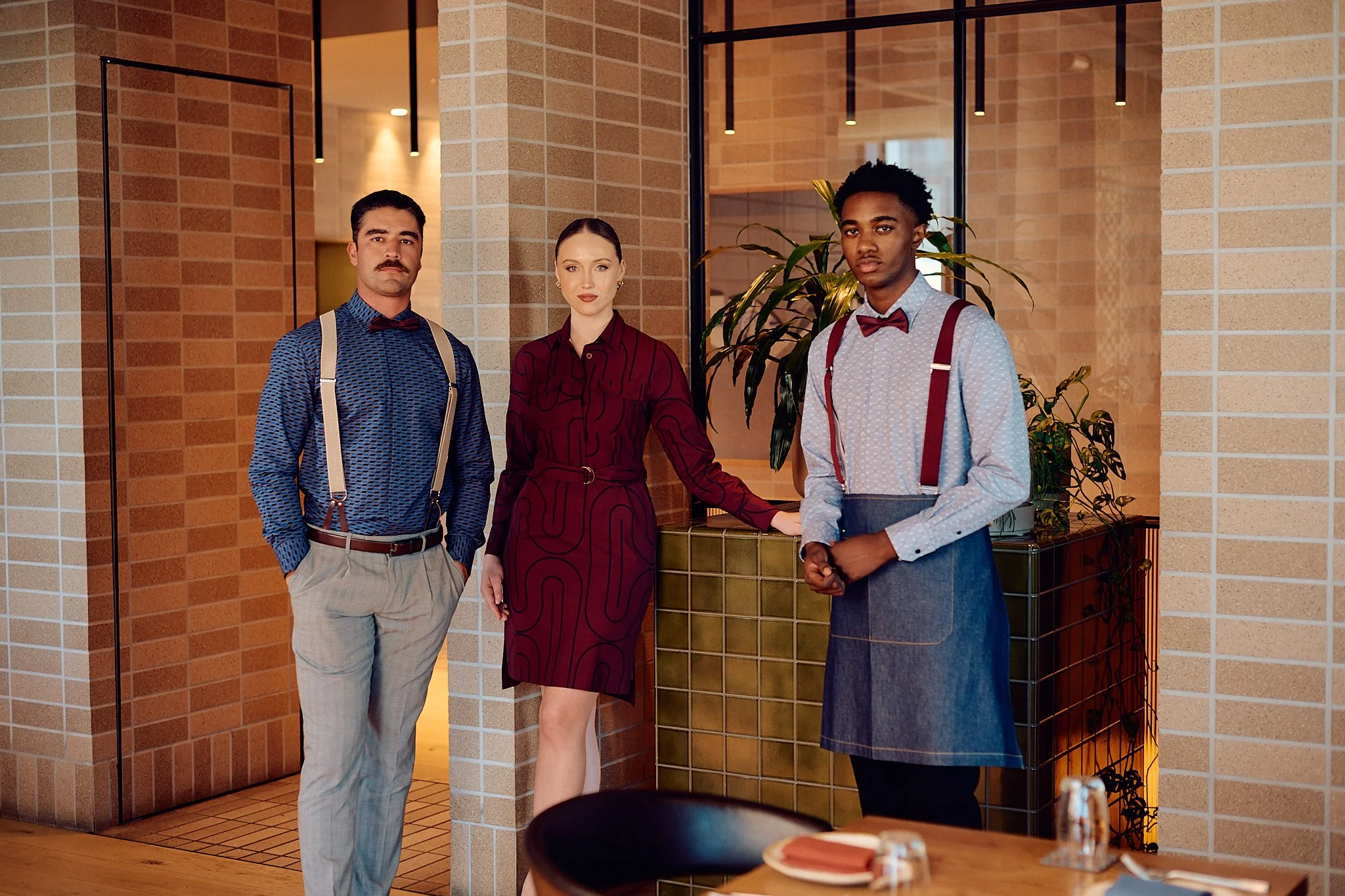 Three waitstaff members standing in a restaurant near a brick wall and a glass window. One man is on the left with dark hair and a mustache, wearing a blue patterned shirt with suspenders. The woman in the middle has blonde hair in an updo, wearing a maroon dress, and the man on the right has short dark hair, wearing a light patterned shirt with suspenders and a dark apron.