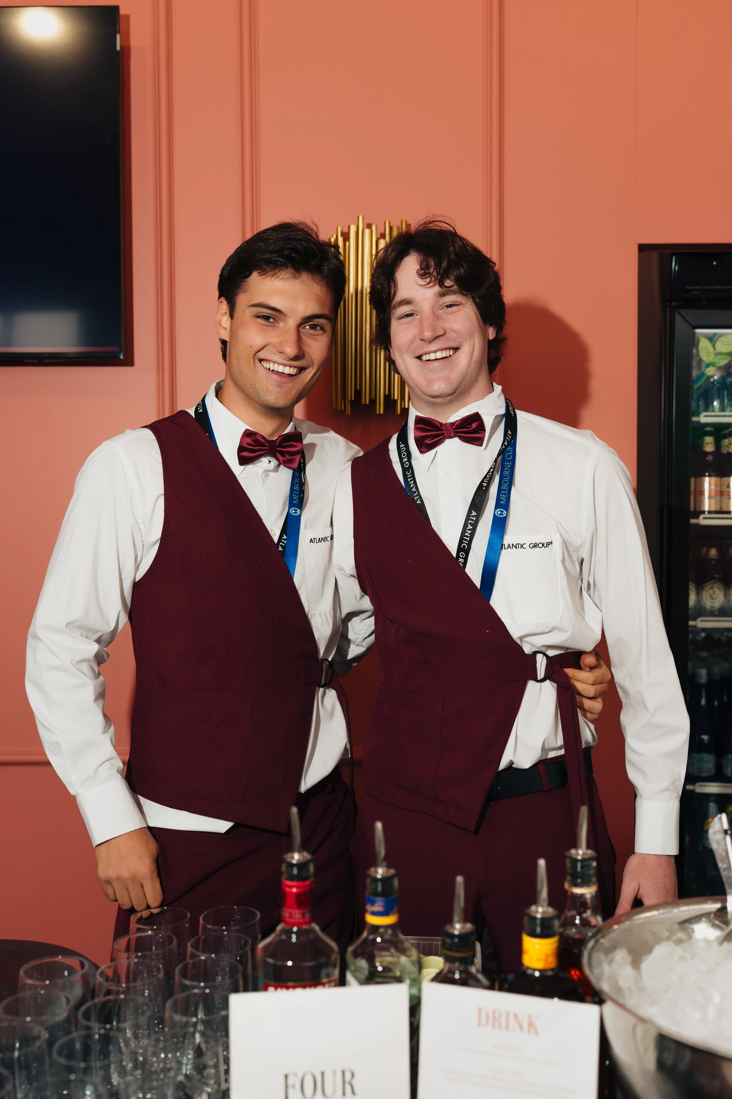 Two smiling male bartenders in white shirts, maroon vests, and bowties behind a bar with liquor bottles and drink glasses.