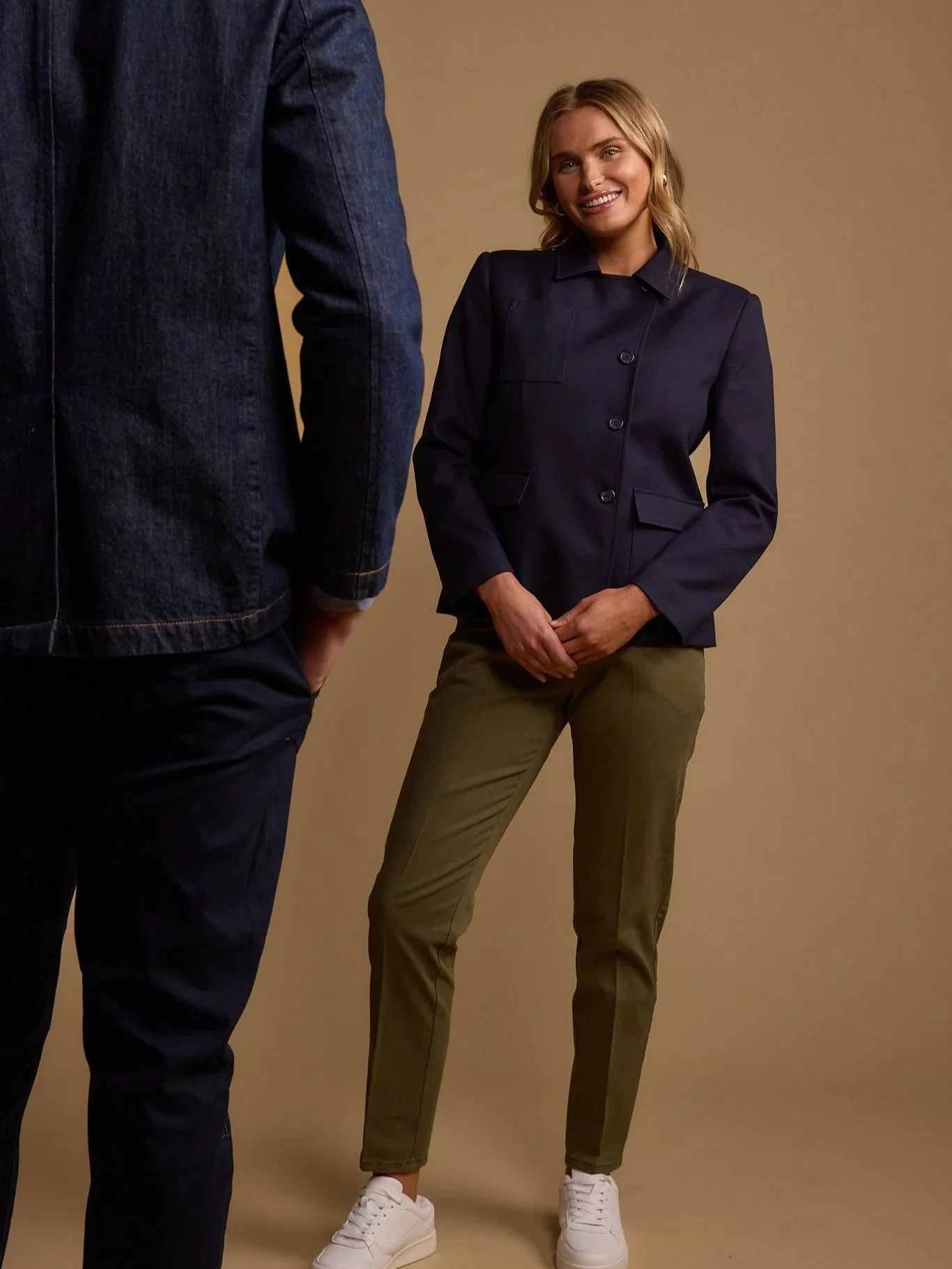 A woman smiling, dressed in a navy blazer, olive green pants, and white sneakers, stands against a beige background. Part of a person wearing a denim jacket and dark pants is visible on the left.
