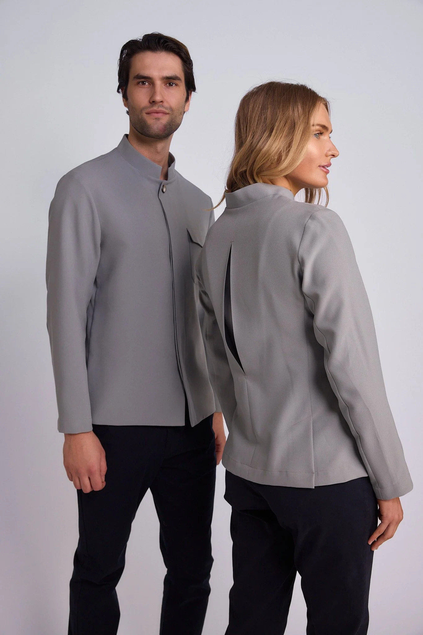 A man and woman in matching light gray jackets and black pants standing against a plain light background. The man faces forward, the woman is turned to the side.