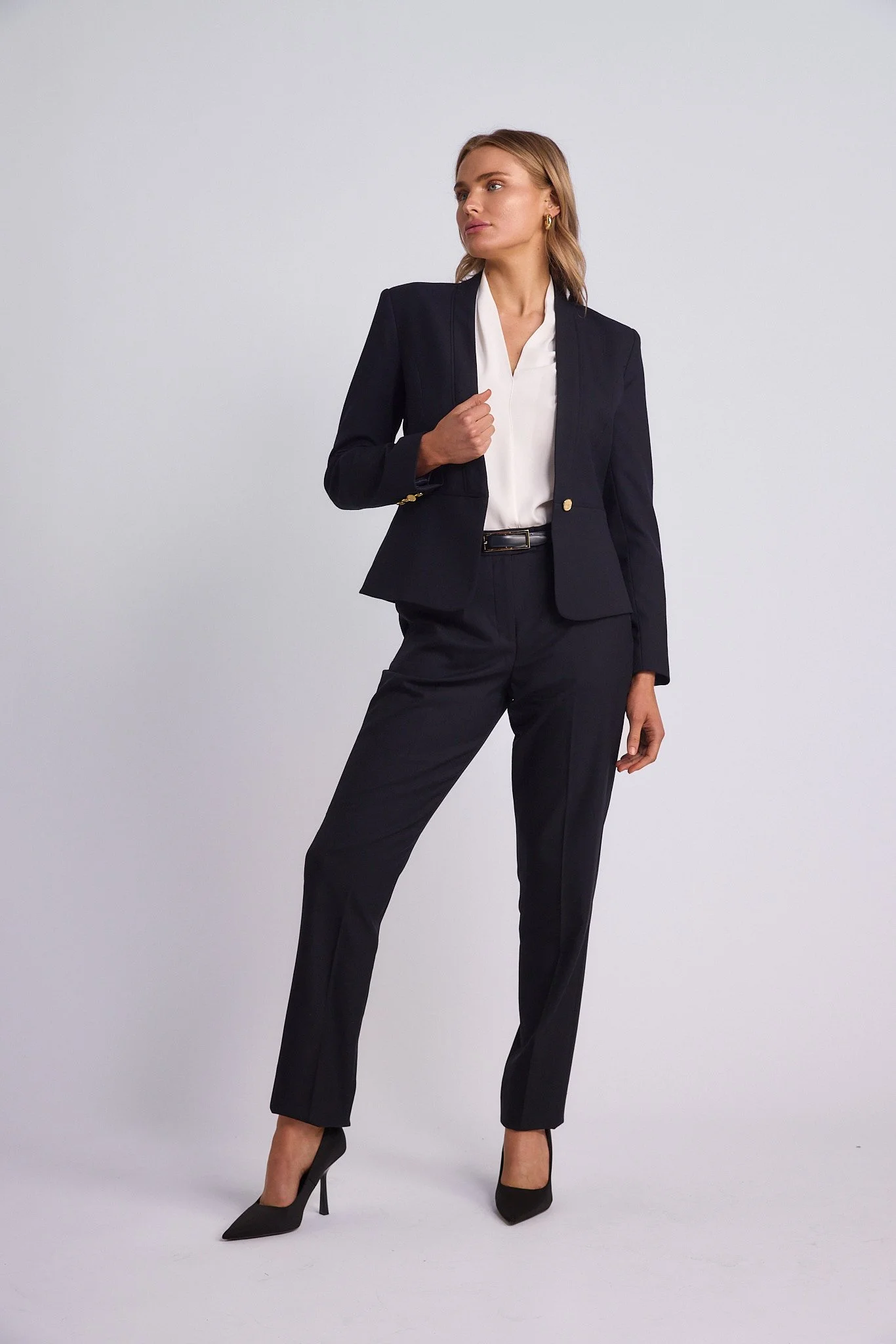 A woman in a dark business suit with high heels, standing confidently against a plain light background.