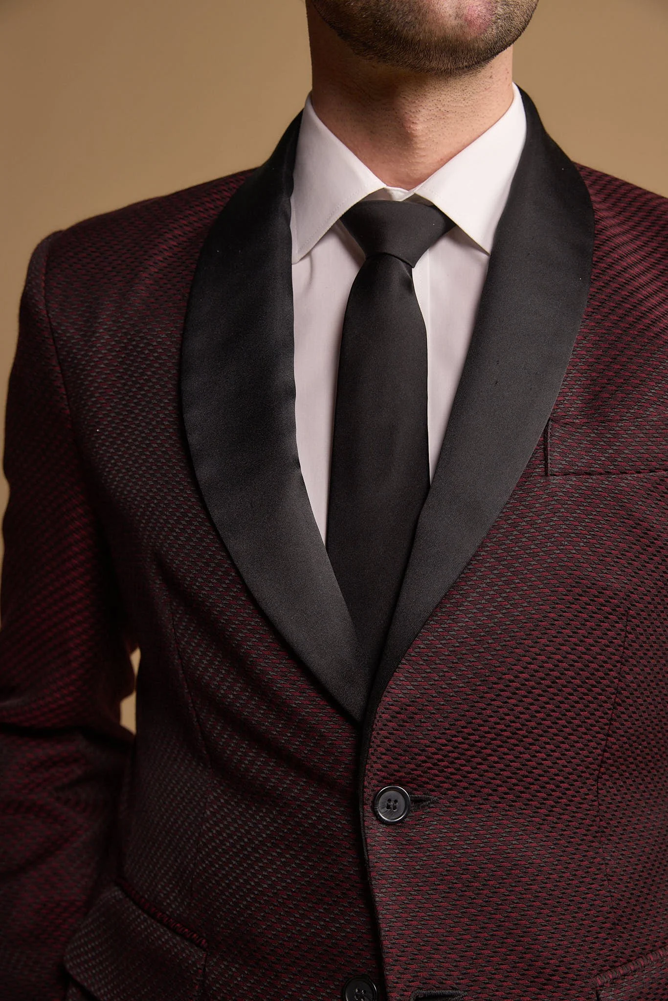 Close-up of a man in a tuxedo jacket with a textured burgundy pattern, a white dress shirt, and a black satin lapel, wearing a black tie against a beige background.