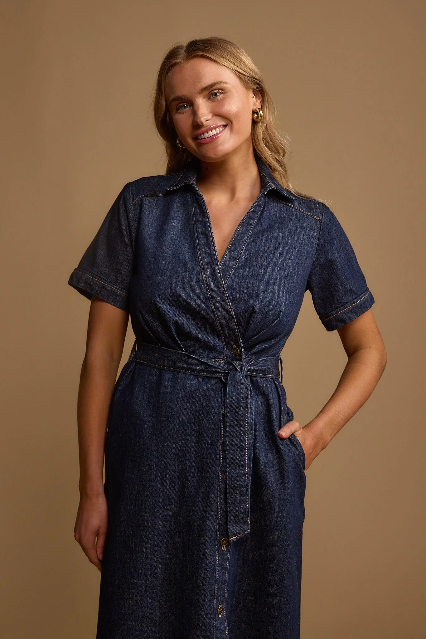 A woman with blonde hair smiling, wearing a dark denim dress with short sleeves and a waist tie, standing against a beige background.
