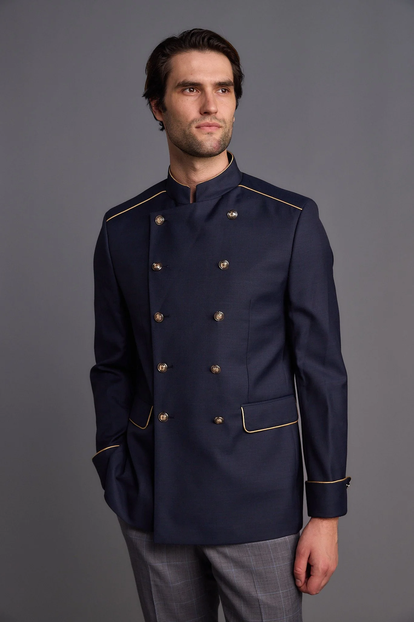 A man in a navy blue double-breasted chef's jacket with gold trim and buttons, standing against a gray background.
