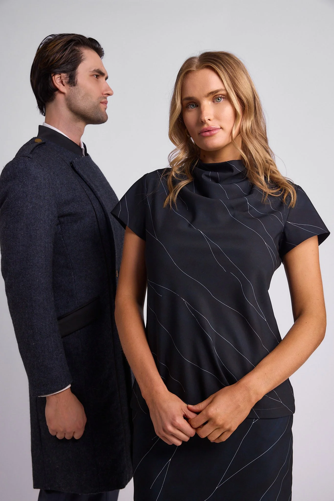 A man dressed as a guard facing a woman wearing a black dress with thin white lines, standing against a plain gray background.