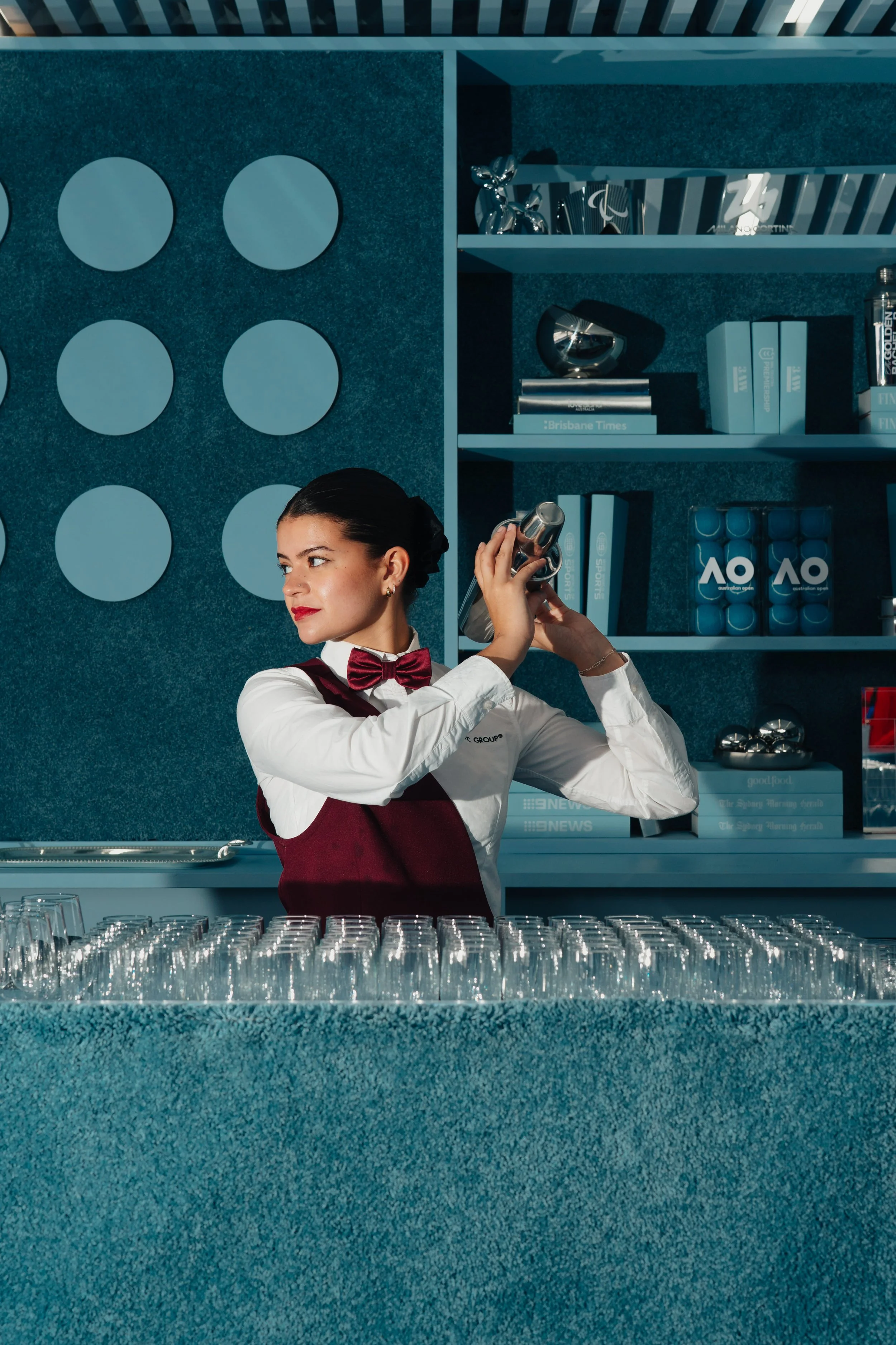 A woman in a formal uniform with a bow tie is shaking a cocktail shaker behind a bar, with multiple glasses arranged on the counter.