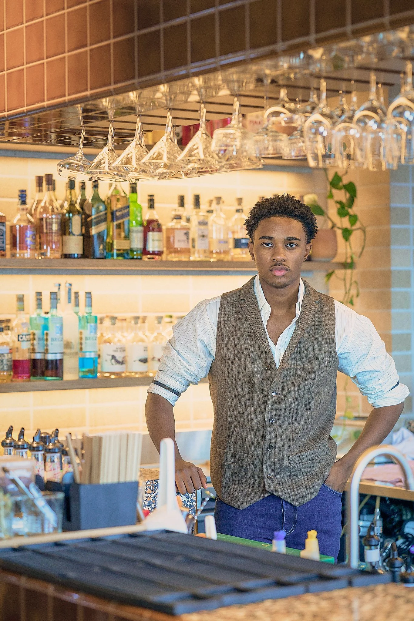A young man standing behind a bar counter in a restaurant or bar, with shelves of liquor bottles and hanging glassware in the background.