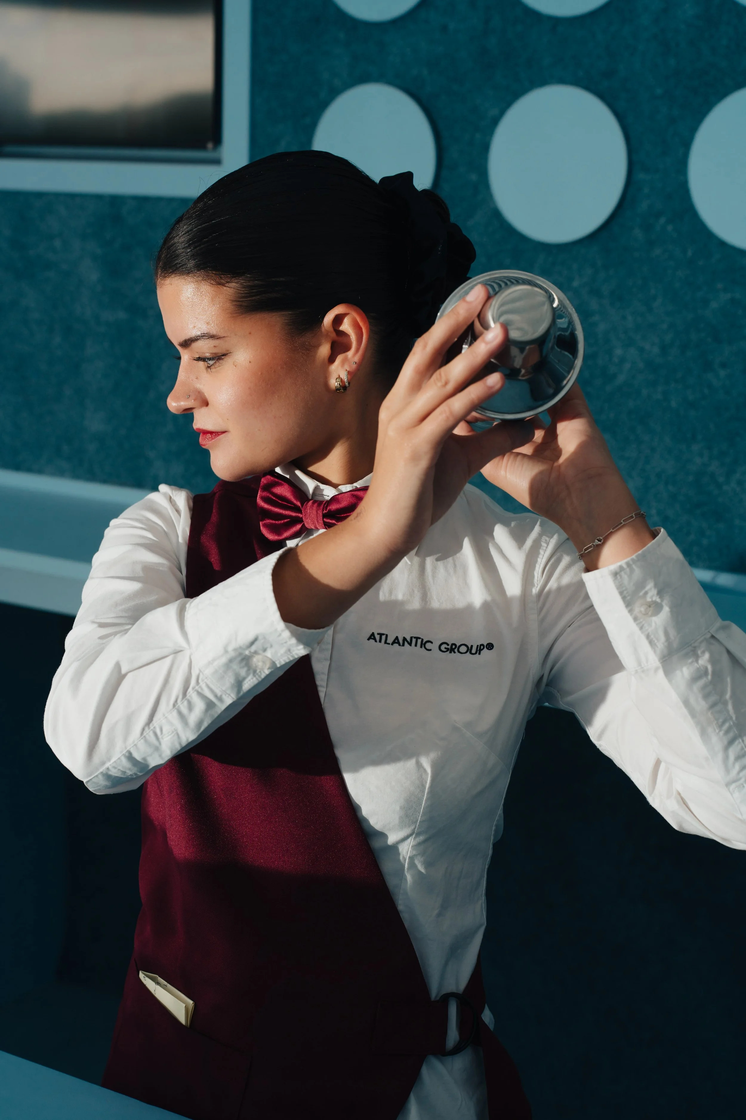 A woman wearing a white shirt with the words 'Atlantic Group' and a red apron, preparing a cocktail in a modern bar or restaurant.