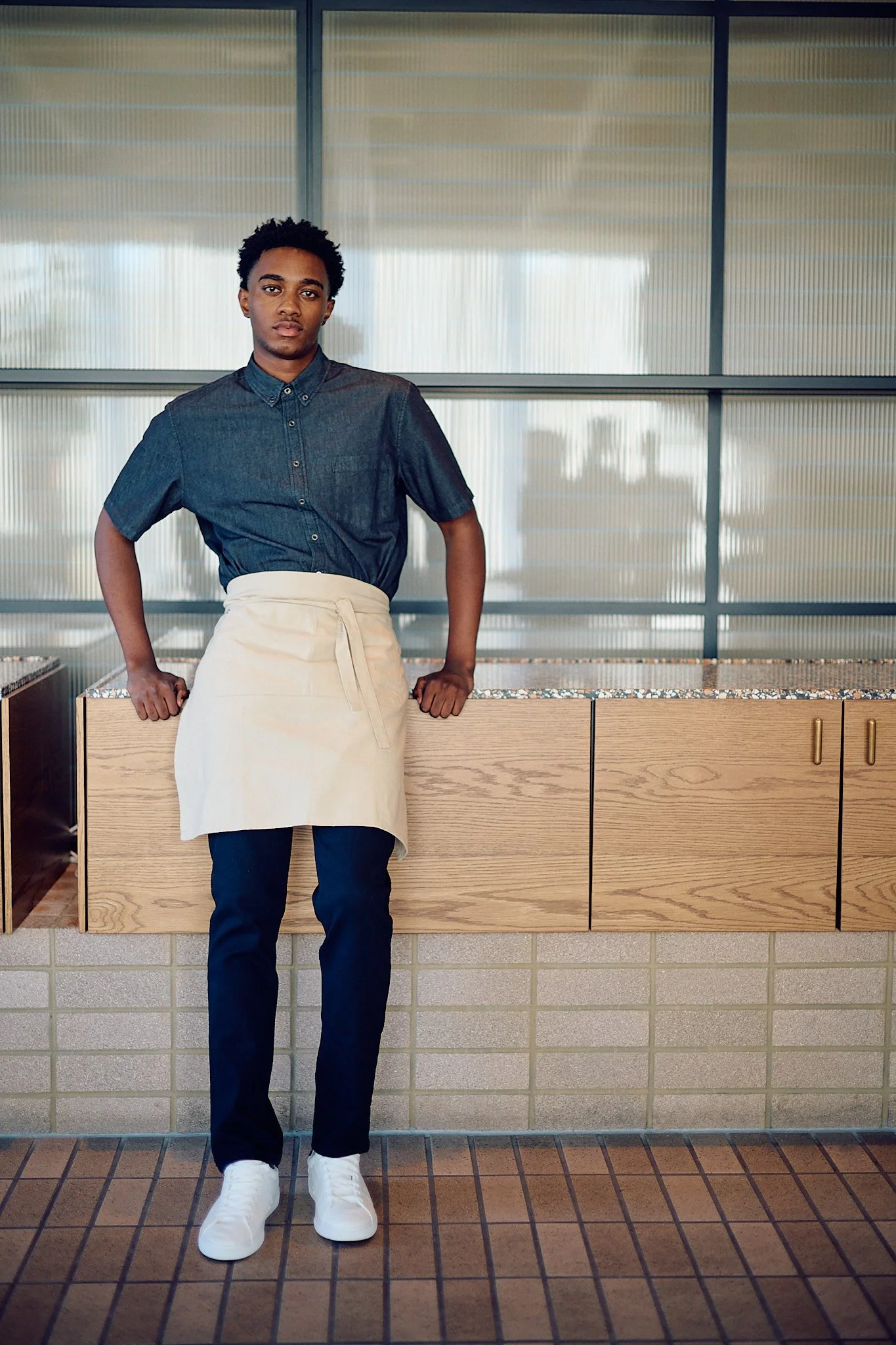 A young man standing indoors in front of a glass window with blinds, wearing a dark denim shirt, white apron, dark pants, and white sneakers.