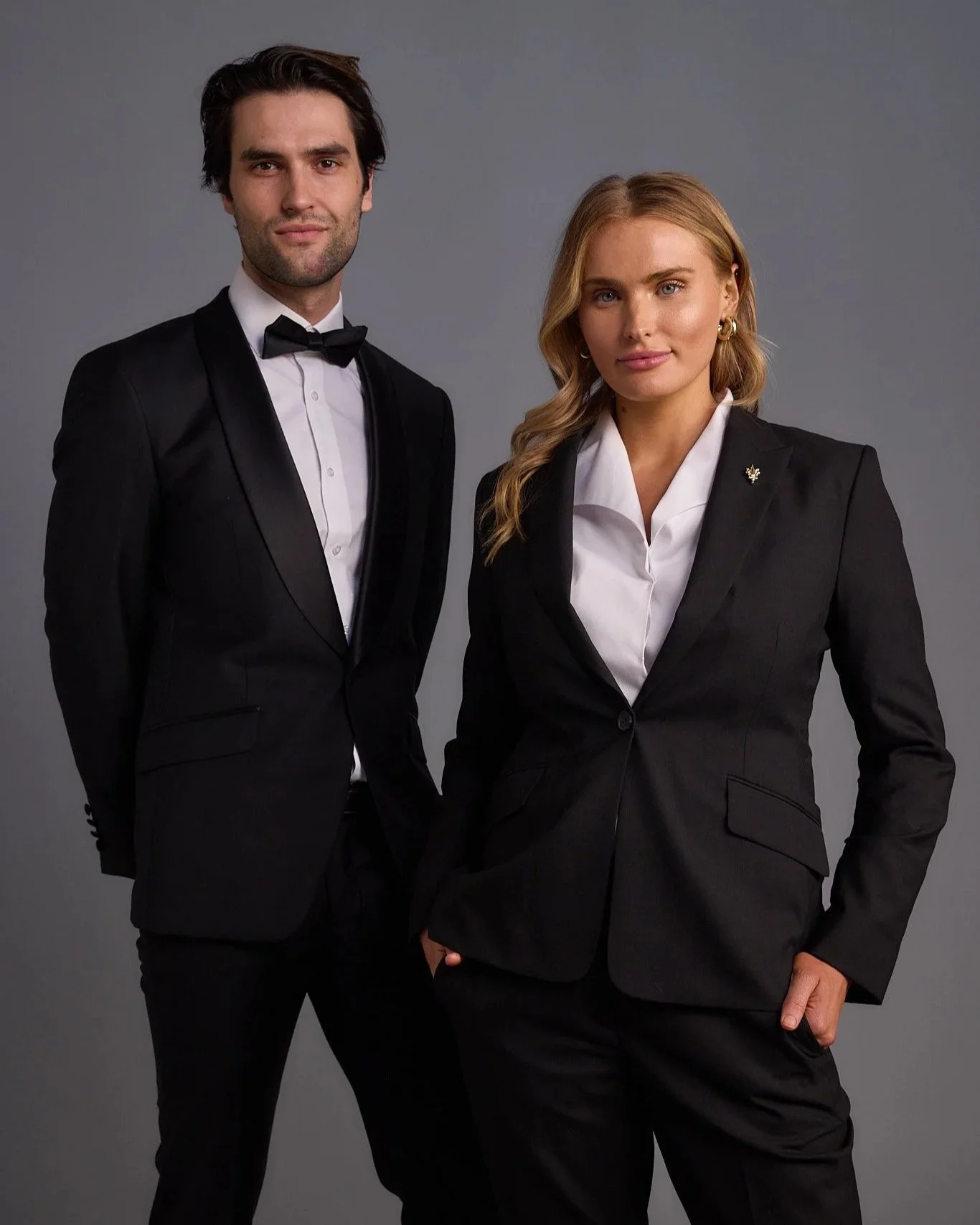 A man and woman dressed in formal black suits, standing side by side against a gray background.