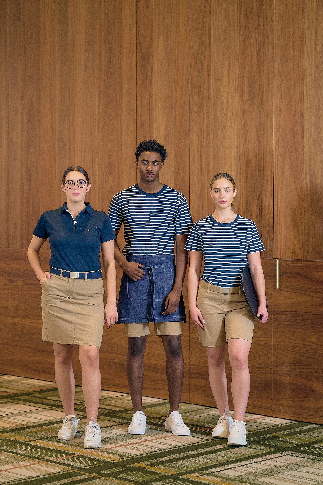 Three young women standing in front of a wooden wall, dressed in similar striped shirts and khaki skirts, with the woman in the center wearing an apron.