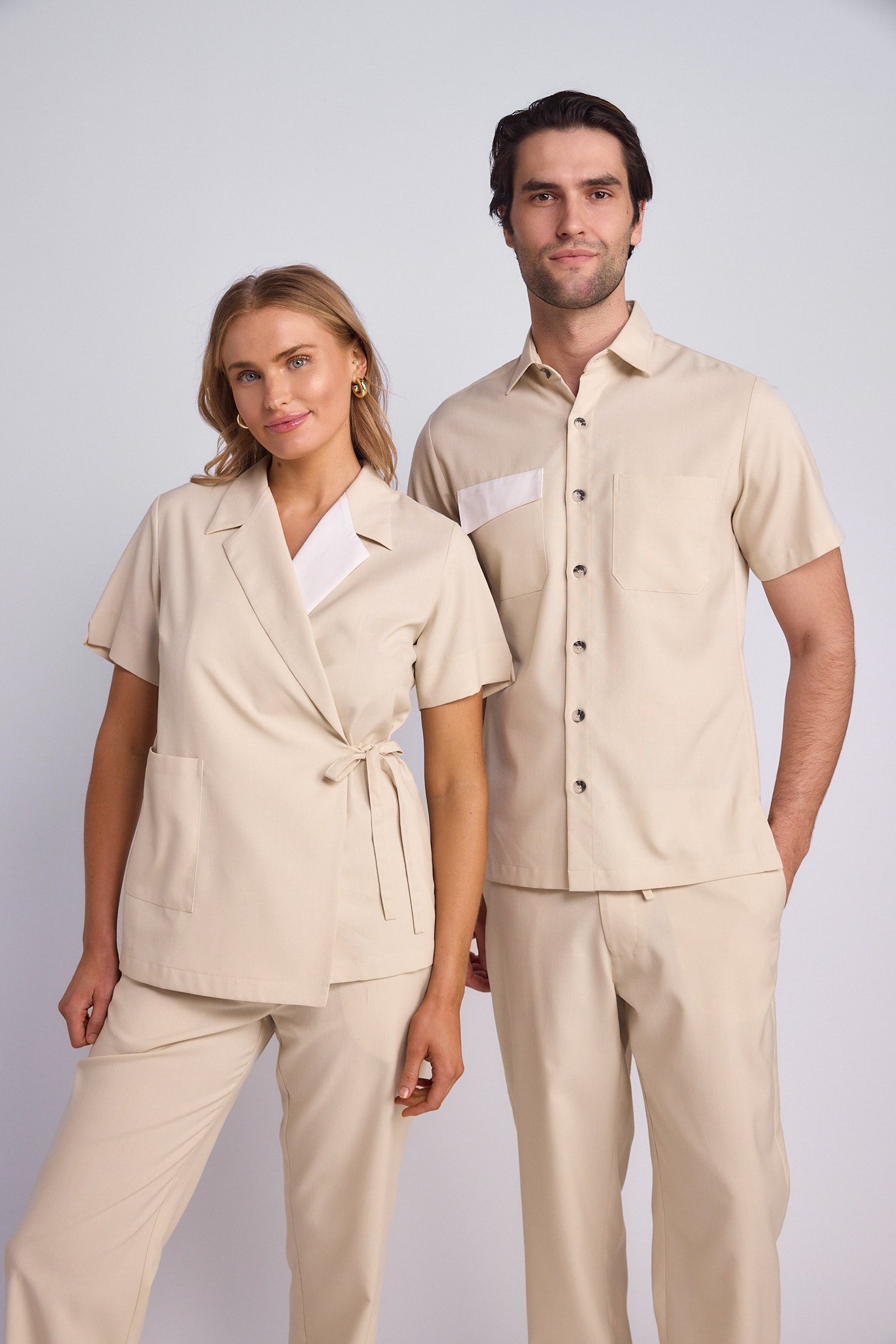 A man and woman dressed in matching beige uniforms, standing side by side against a light gray background.
