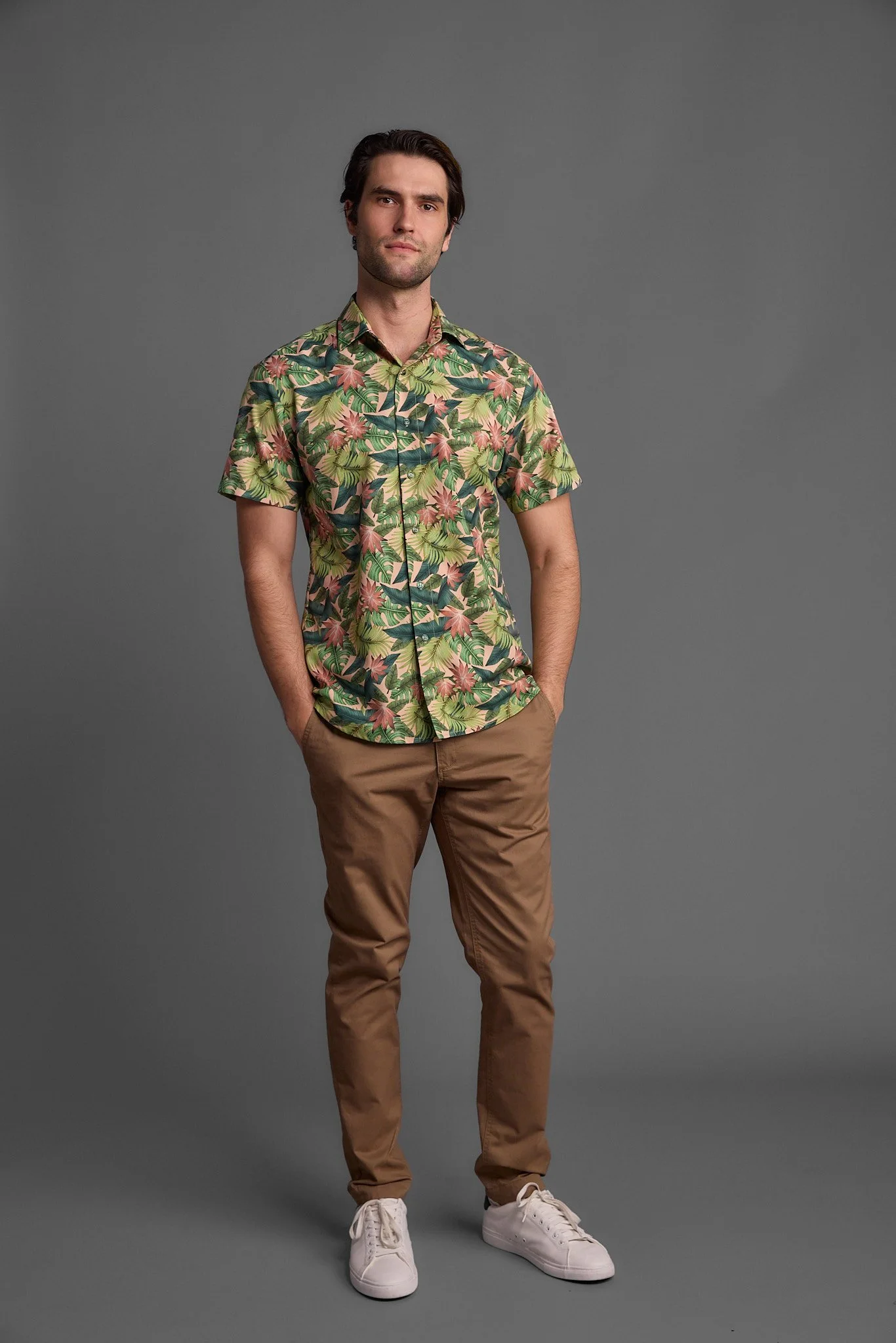 Young man standing against a gray background, wearing a短-sleeve tropical shirt, tan pants, and white sneakers.