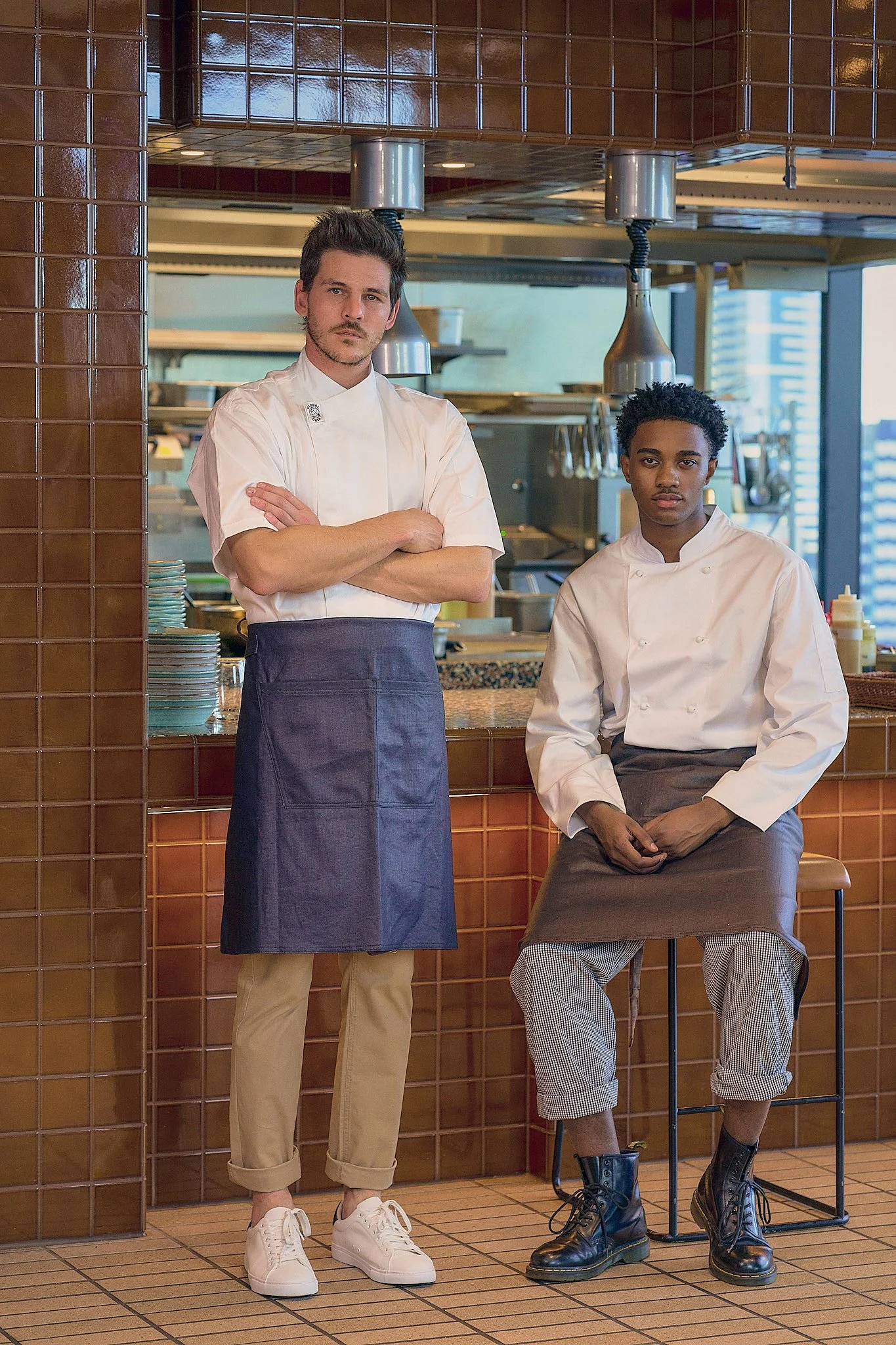 Two chefs in white uniforms, one standing with arms crossed and the other sitting on a stool, in a restaurant kitchen with brown tiled walls and stainless steel cooking equipment.