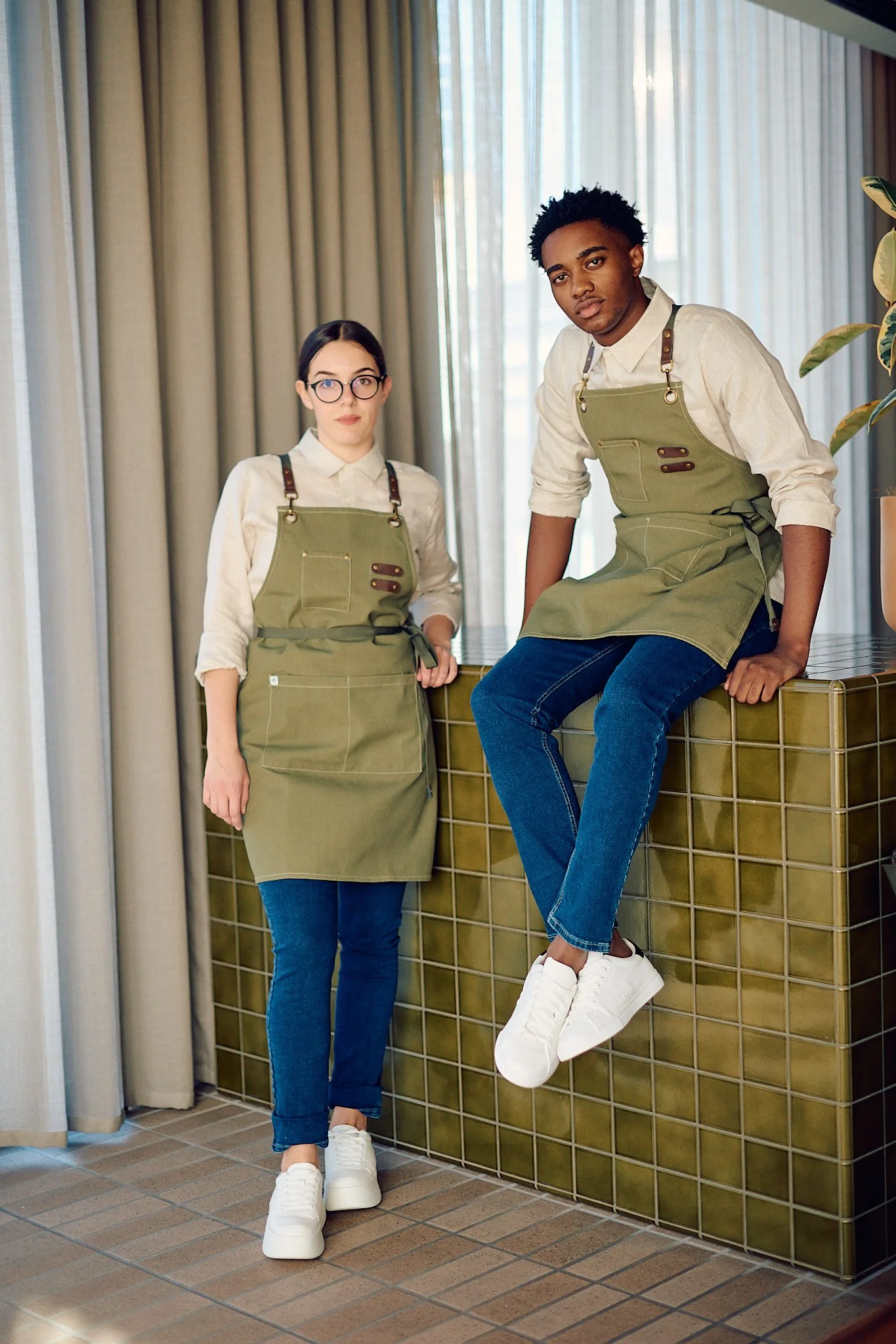 Two young people, a woman and a man, both wearing beige shirts, green aprons, and white sneakers, standing indoors near a tiled counter with large windows and curtains in the background.
