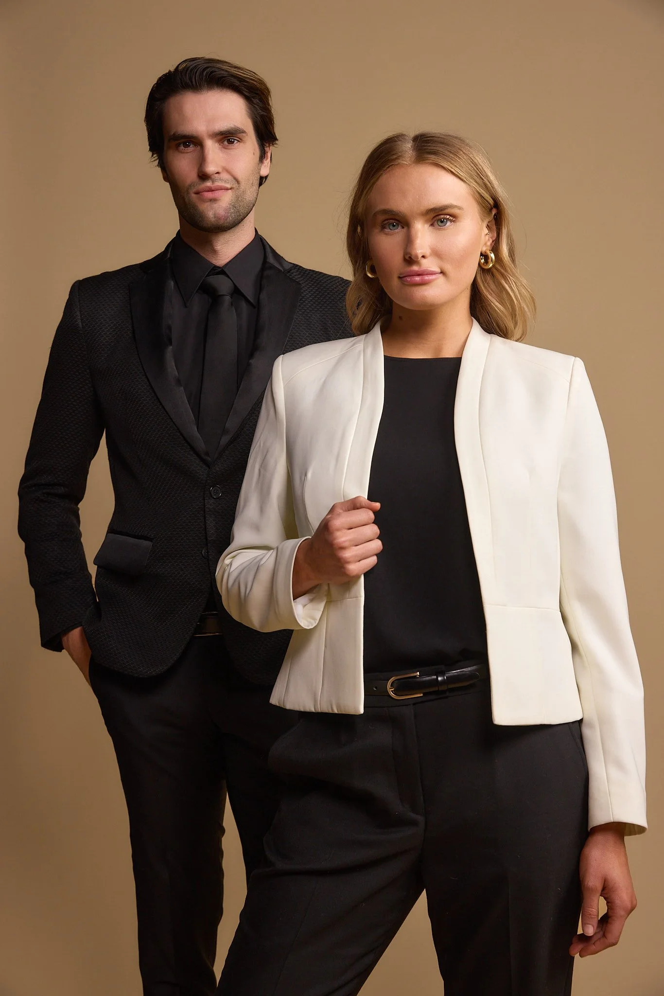 A man and woman in formal business attire posing against a beige background. The man is dressed in a black suit and black shirt, standing slightly behind and to the left of the woman. The woman wears a white blazer over a black top and black trousers