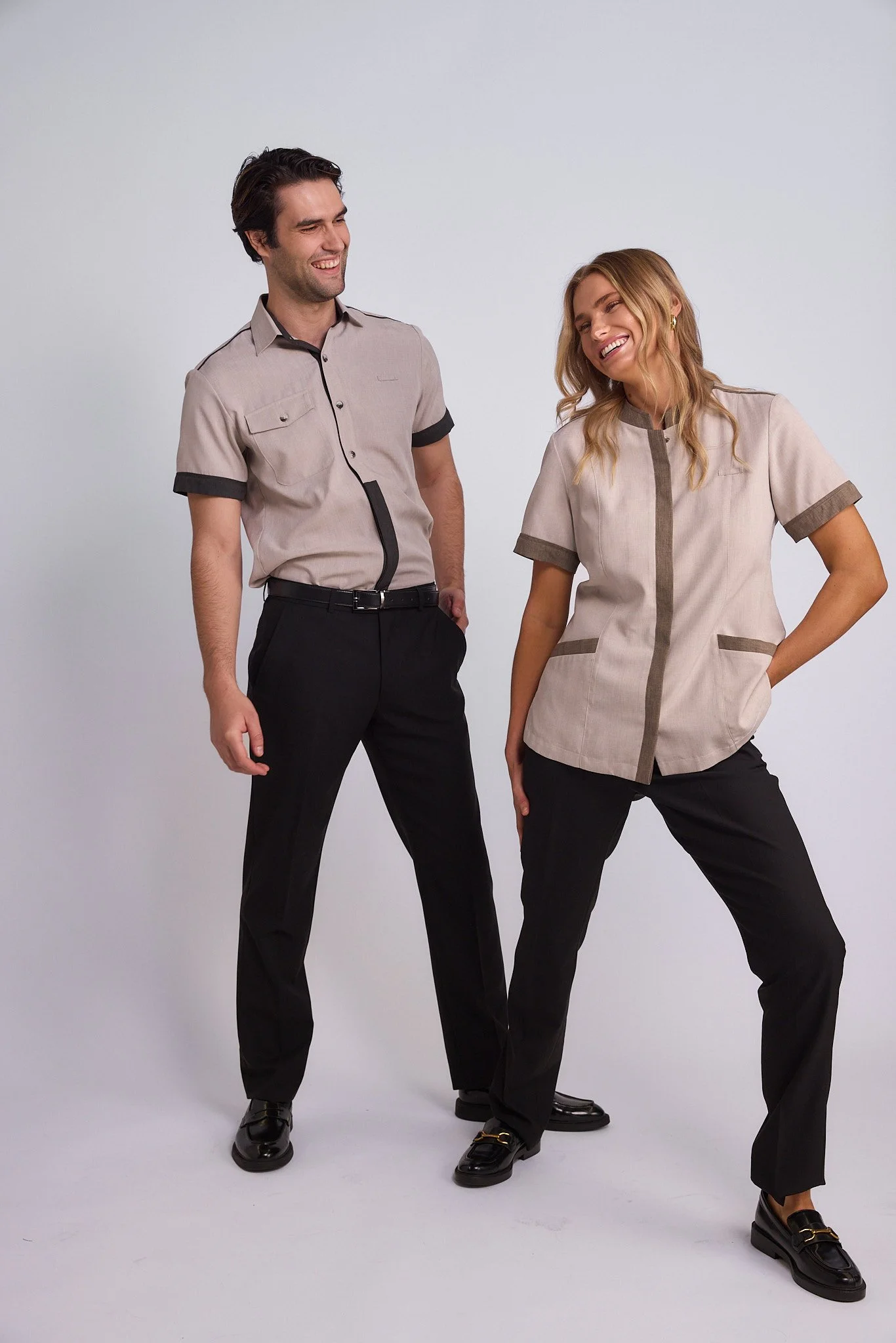 Two people, one man and one woman, standing and laughing together. They are wearing coordinated beige and black outfits and black shoes. The background is plain and white.