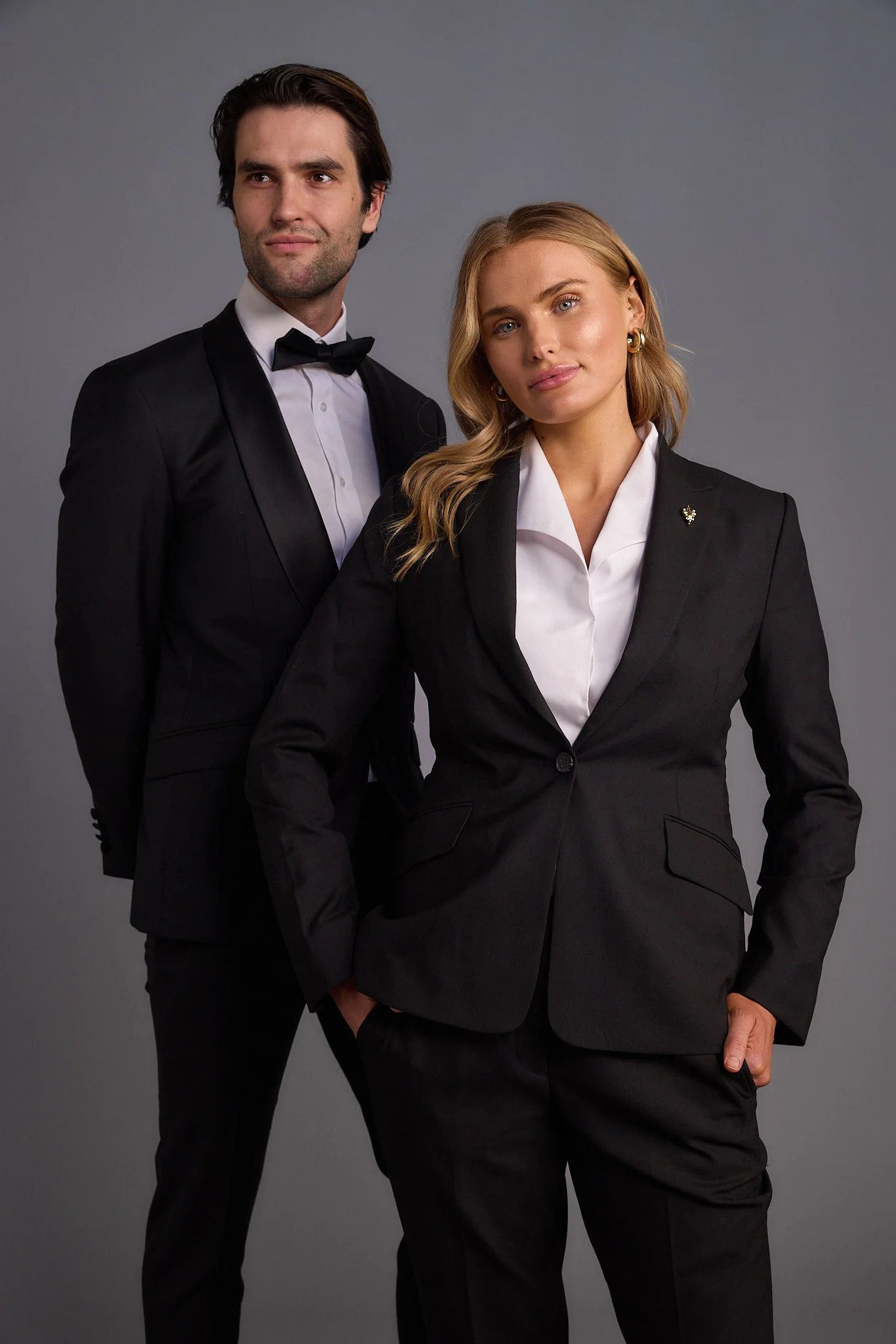 A man and woman dressed in black business suits with white shirts, standing against a gray background. The man is in the back, wearing a tuxedo with a bow tie, and the woman is in the front with her hand in her pocket, wearing a blazer and matching p