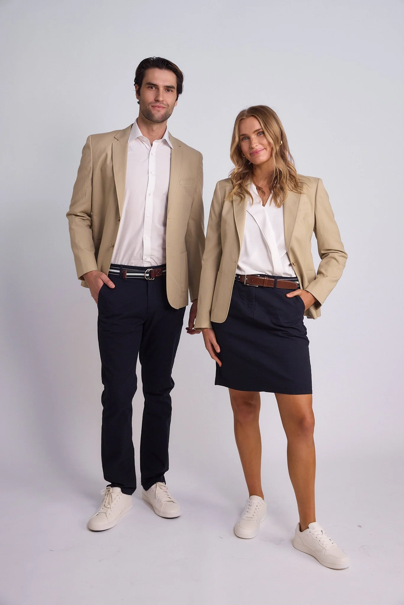 Two people, a man and a woman, standing side by side against a plain white background. They are dressed in business casual attire with beige blazers, white shirts, navy pants or skirt, and white sneakers.