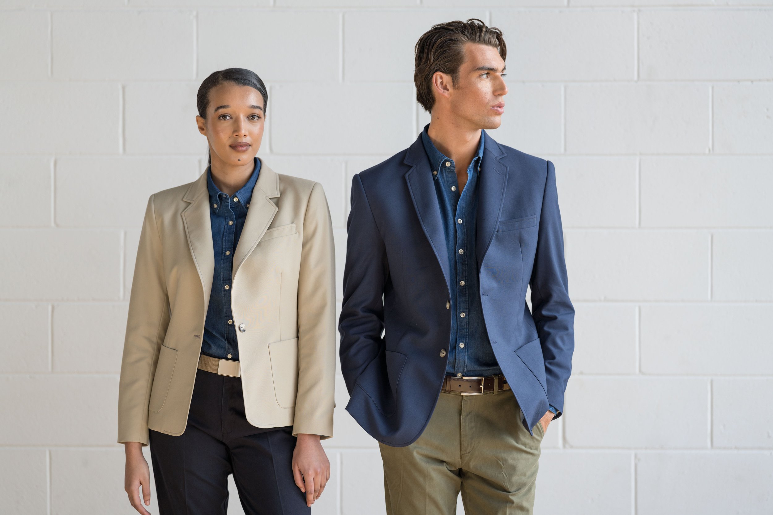 A woman and a man standing side by side against a white brick wall. The woman is wearing a beige blazer over a denim shirt with black pants. The man is wearing a navy blazer over a denim shirt with khaki pants.