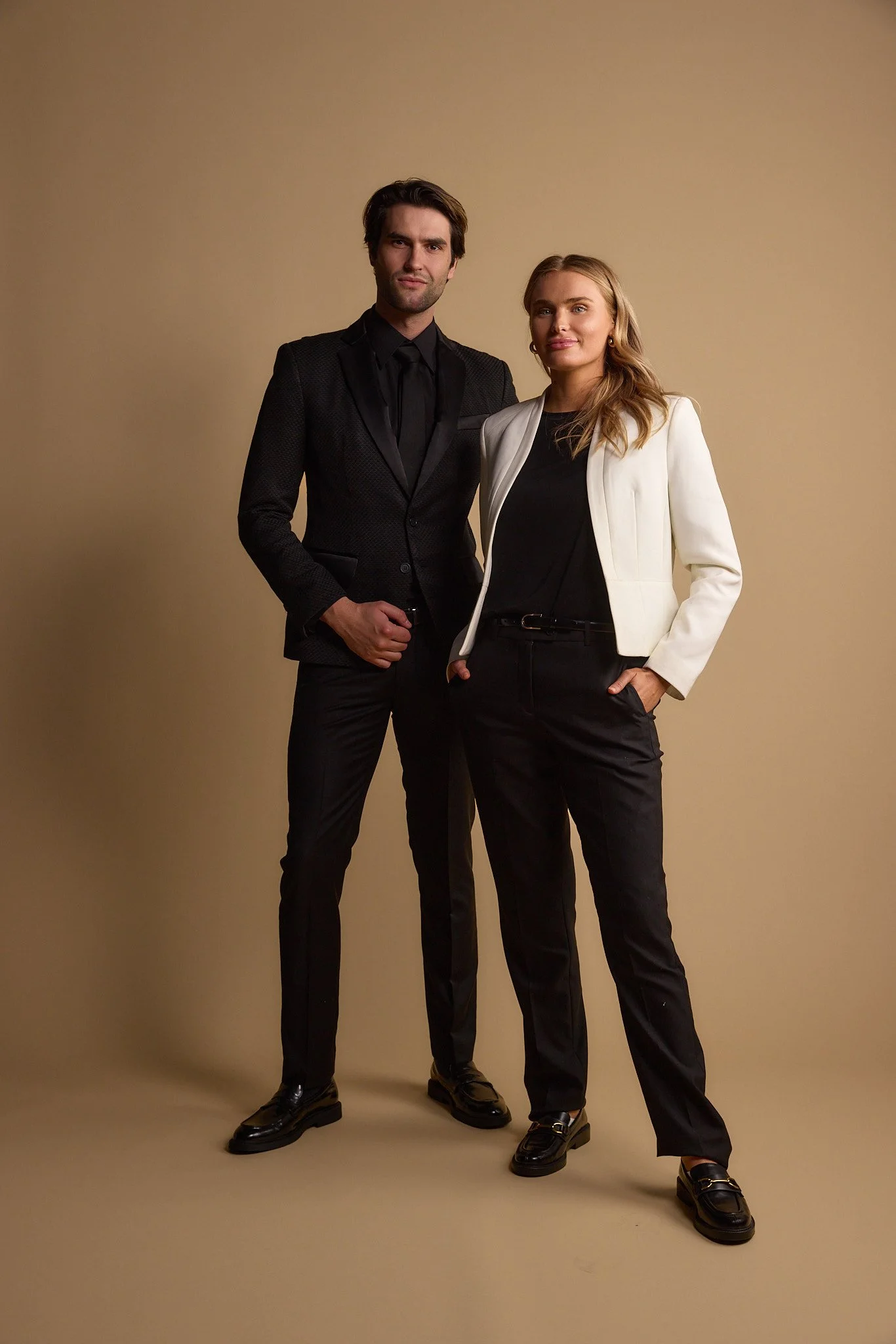 A man and woman dressed in formal business attire standing against a beige background.