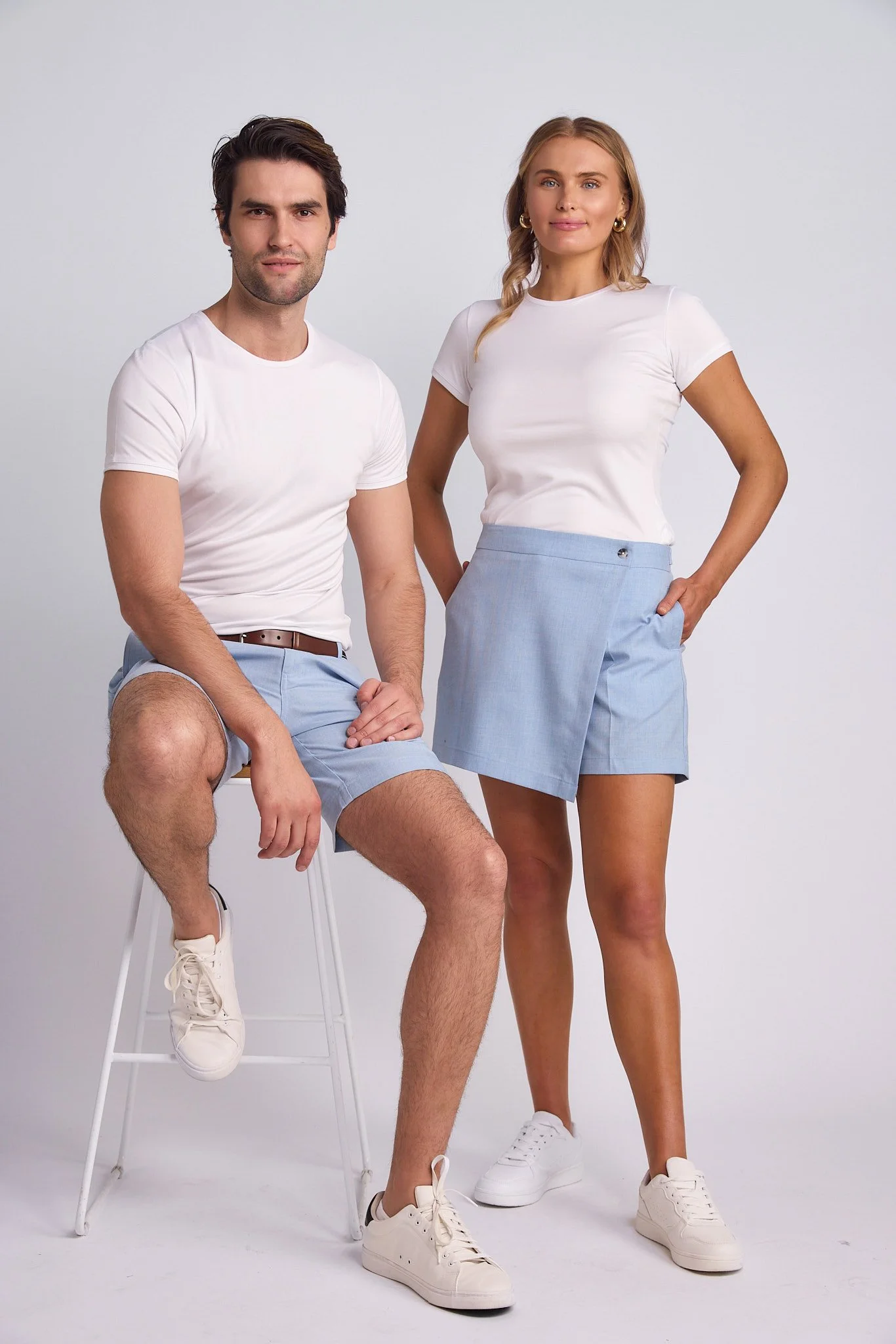 A man and woman wearing white t-shirts, light blue shorts, and white sneakers, posing against a plain white background.