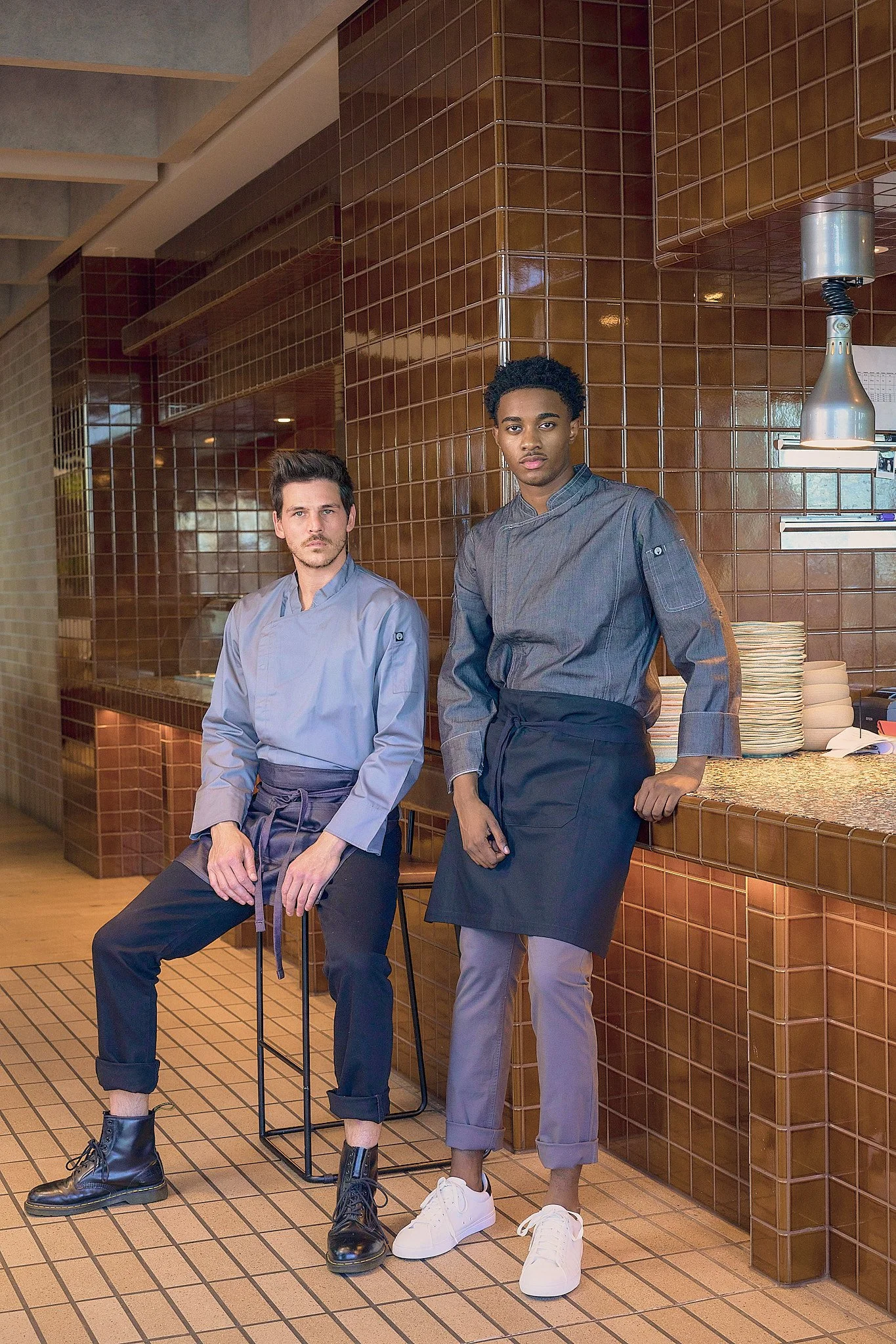Two chefs in gray uniforms inside a restaurant kitchen, one is sitting on a stool and the other is leaning against the counter, both looking at the camera.