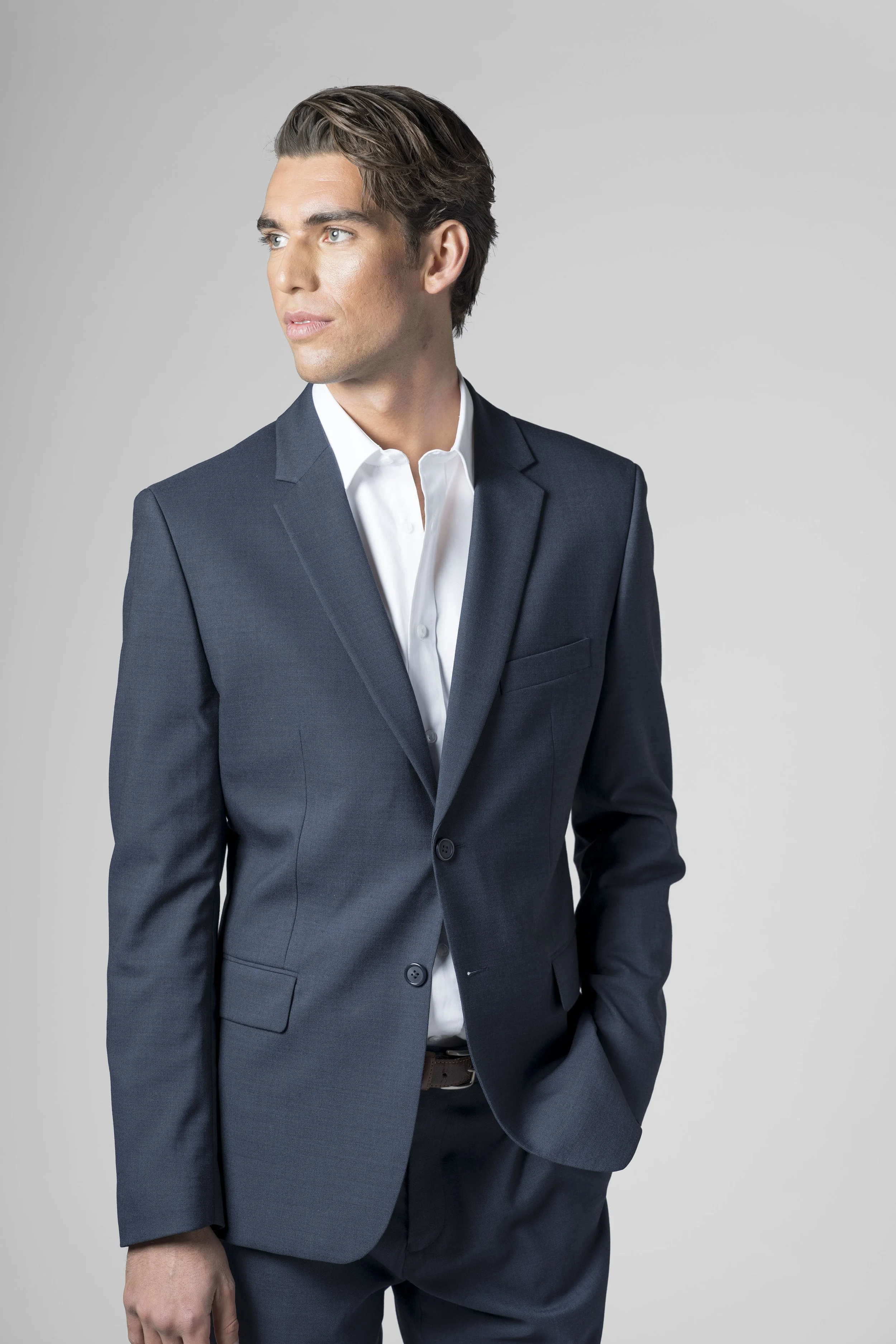 A man in a dark suit with a white shirt standing against a plain gray background.