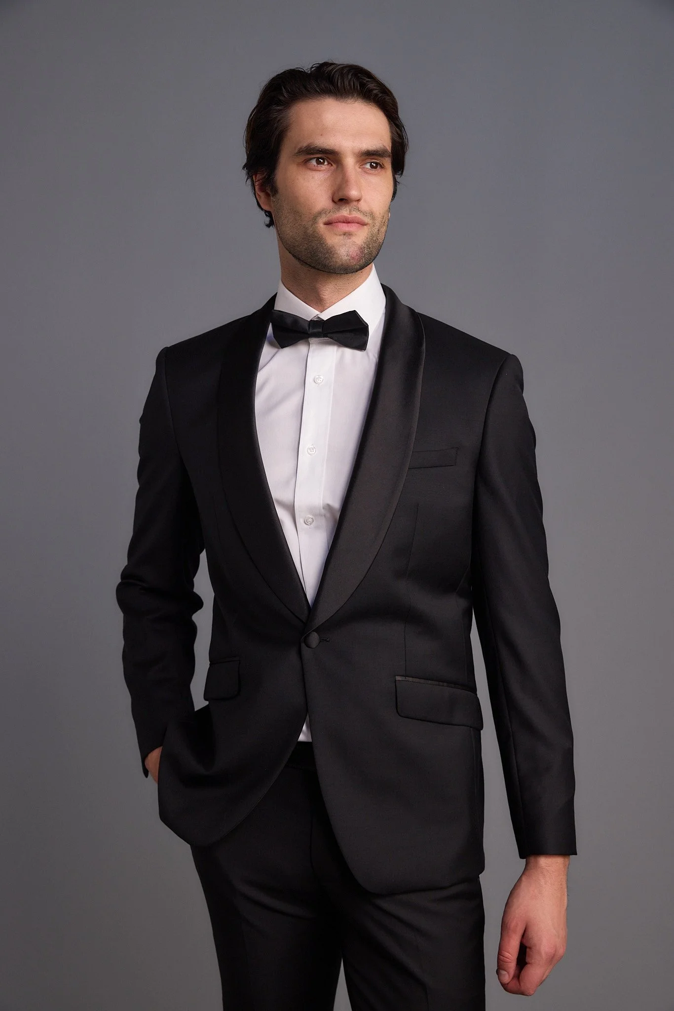 A man in a black tuxedo with a white shirt and black bow tie posing against a plain gray background.
